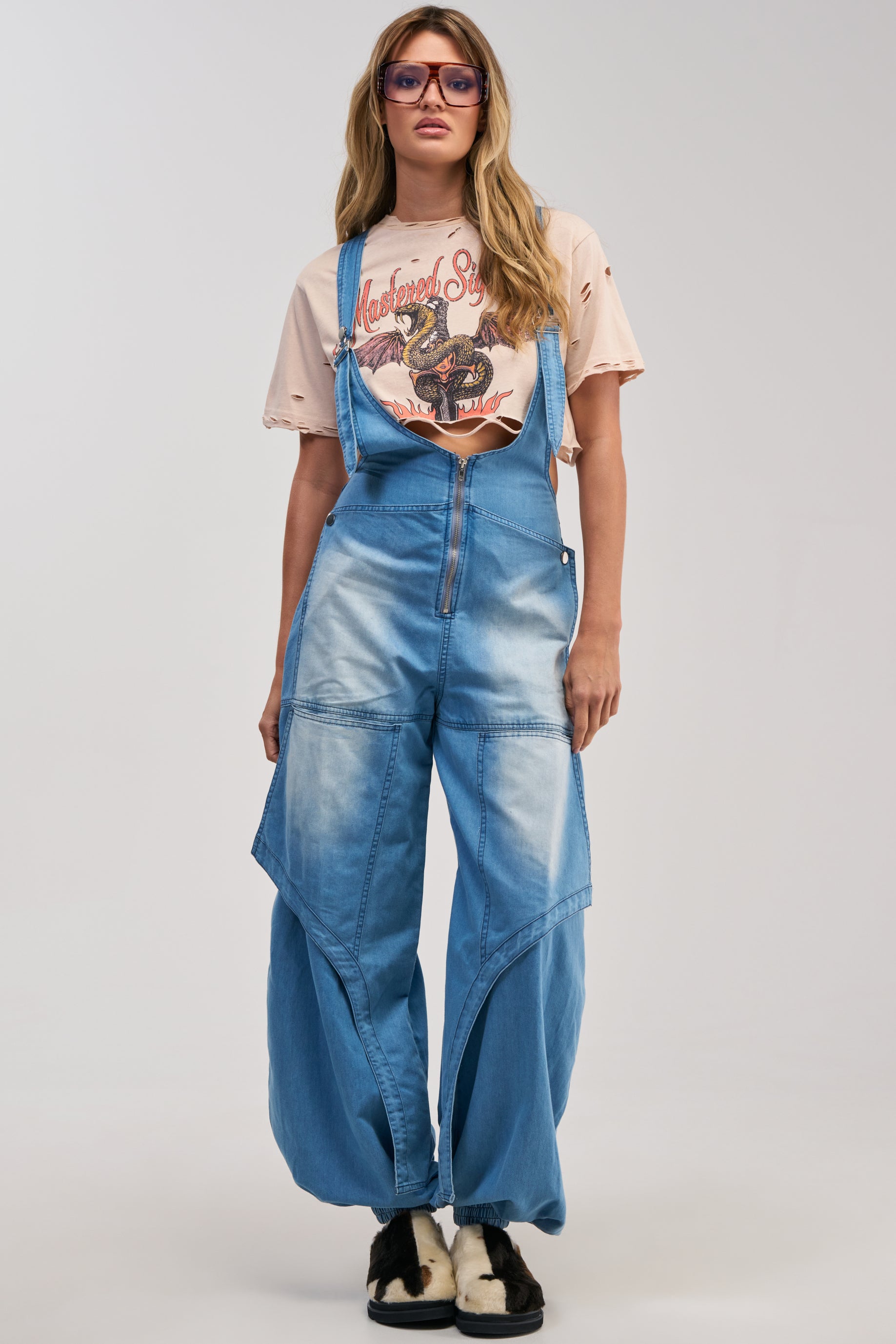 A person wearing the OUT TO PLAY DENIM OVERALL JUMPSUIT, styled with a graphic T-shirt, black-and-white shoes, and purple sunglasses, stands against a plain background.