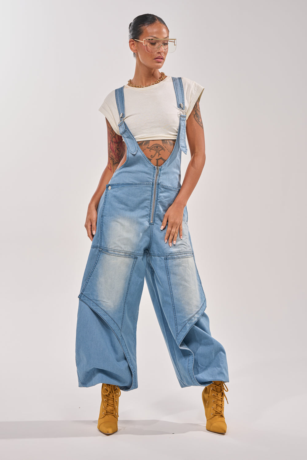 A person confidently wears the OUT TO PLAY DENIM OVERALL JUMPSUIT with wide legs and a front zipper, styled with a white cropped tee, yellow heeled boots, clear glasses, jewelry, tattoos, and a sleek hairstyle against a plain light background. 1