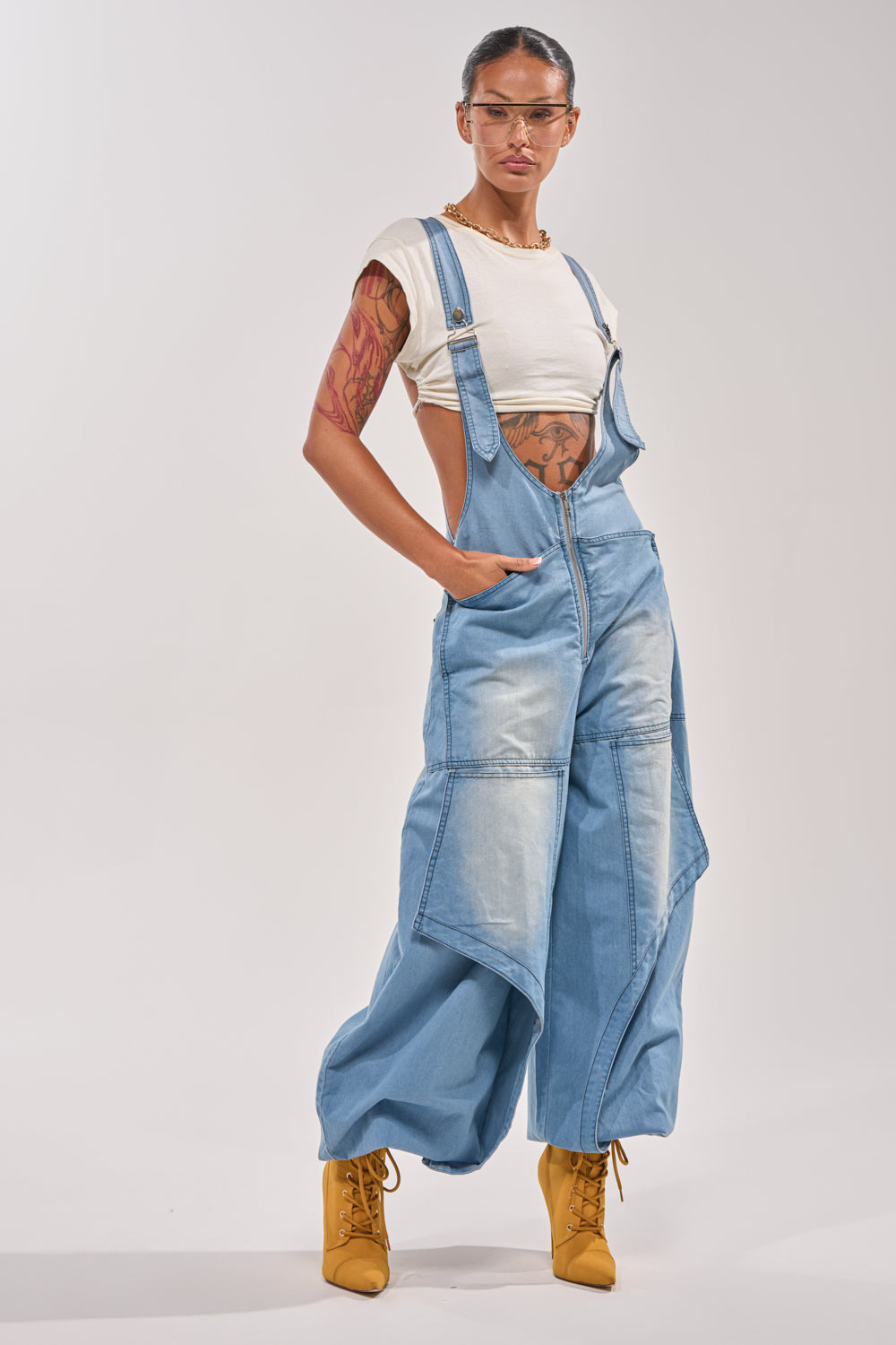 A person in the OUT TO PLAY DENIM OVERALL JUMPSUIT, white cropped tee, yellow lace-up boots, and clear glasses poses with hands in pockets against a plain light background, showing tattoos on their arms. 2