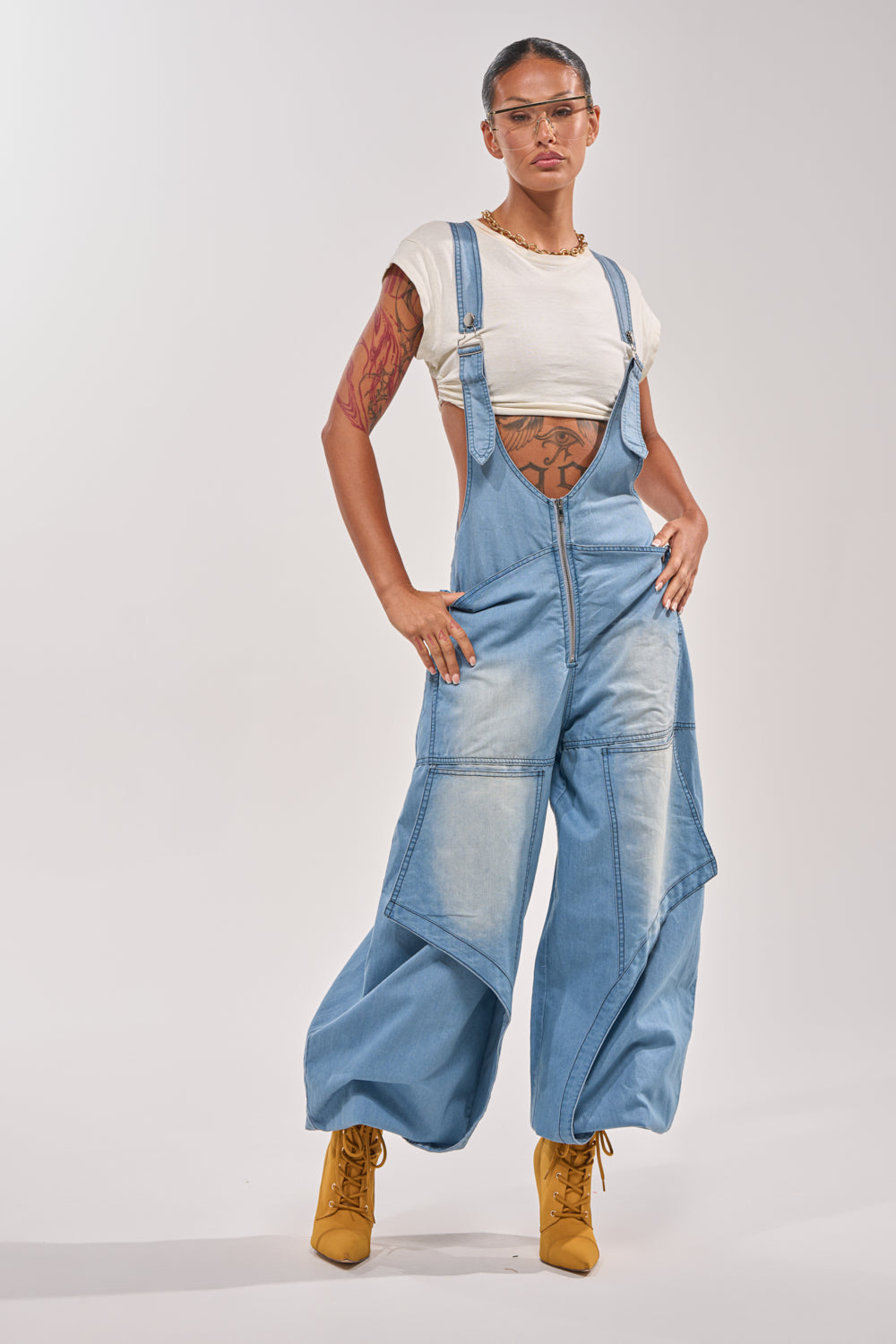 A person wearing the OUT TO PLAY DENIM OVERALL JUMPSUIT, a white cropped tee, yellow boots, clear glasses, and jewelry stands confidently with tattooed arms and a neutral expression against a plain background. 3