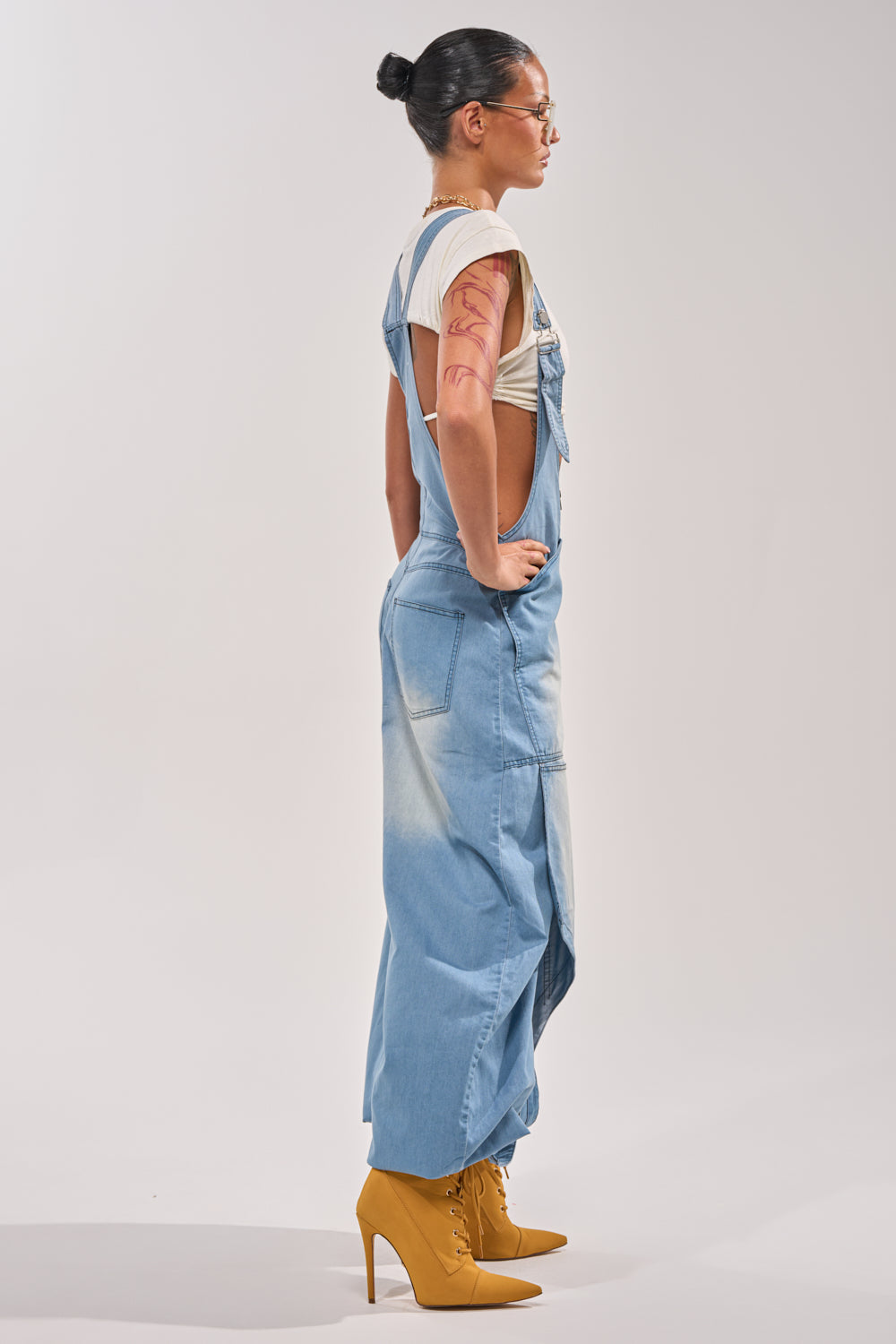 A person with dark hair in a bun, glasses, a white T-shirt, and yellow high-heeled boots models the OUT TO PLAY DENIM OVERALL JUMPSUIT in light blue against a plain background. 4
