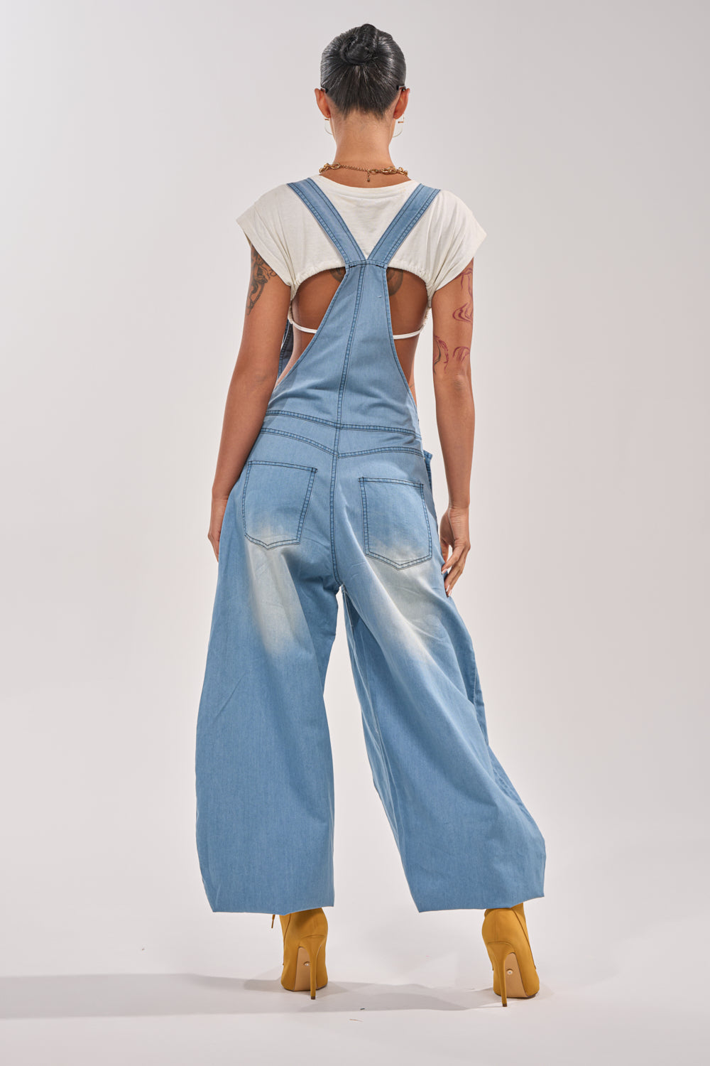 A person with short dark hair stands facing away, wearing the OUT TO PLAY DENIM OVERALL JUMPSUIT with wide legs over a white short-sleeve shirt and yellow high-heeled shoes, set against a plain light background. 5