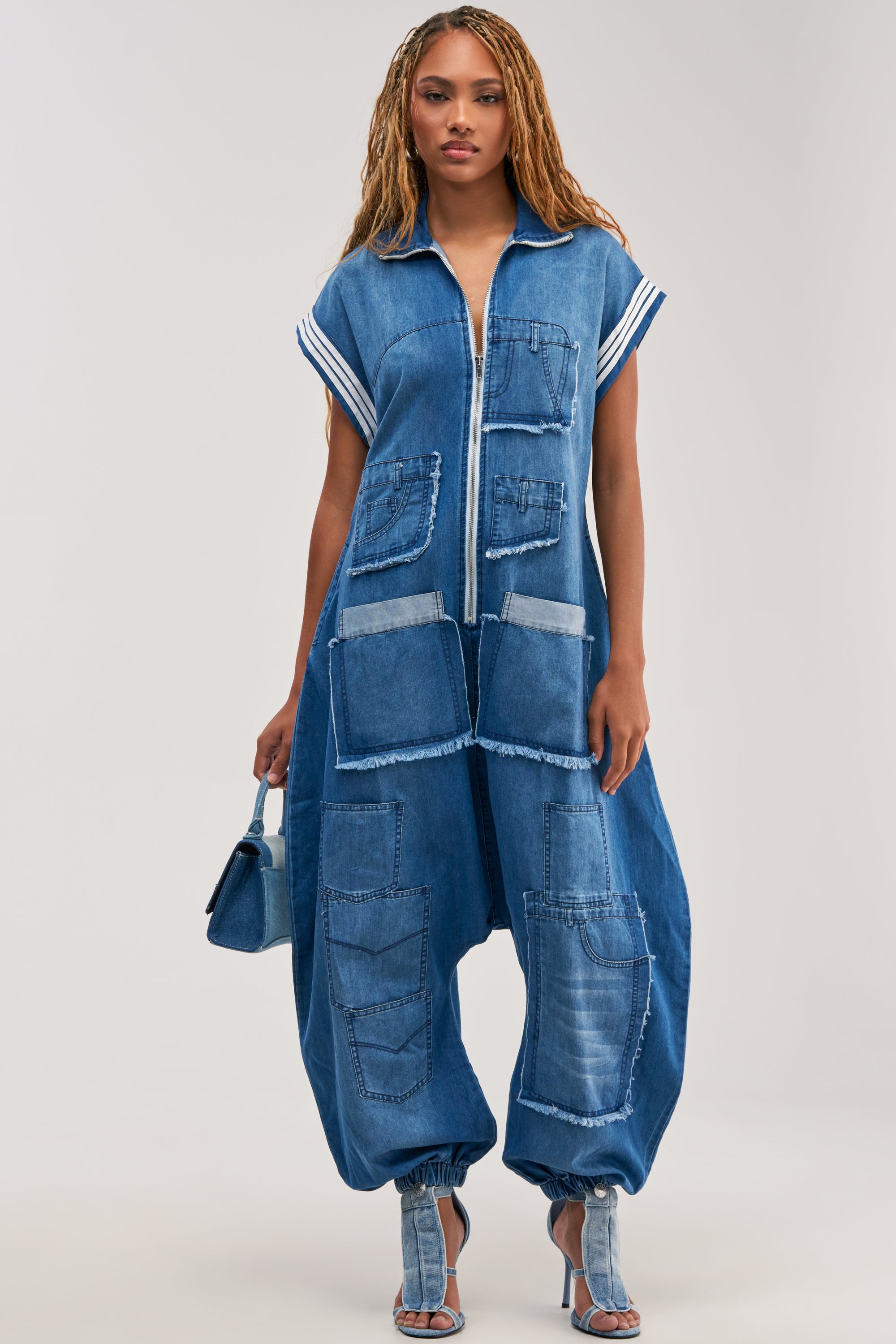 A person stands against a plain background, effortlessly channeling modern street style in the OFF-DUTY DENIM PATCHWORK JUMPSUIT—featuring a loose silhouette, multiple pockets, patchwork details, cuffed ankles—styled with open-toe heels and a small denim bag.