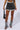 A person is wearing the JCR Trading LTD GET IT RIGHT DISTRESSED DENIM PLEATED MINI SKIRT over white shorts, a cropped top, and denim leg warmers with sandals. They are holding a black handbag with their left hand. Part of the Get It Right Collection, the outfit is trendy and stylish, mixing denim and casual elements effortlessly.