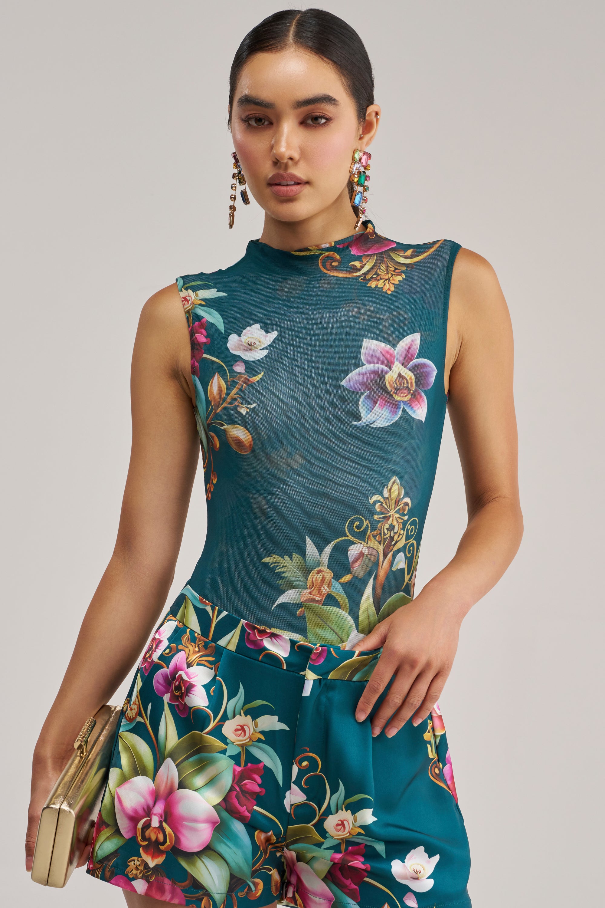 A woman wearing the ISLAND TIME SLEEVELESS MESH BODYSUIT, a high-neck, sleeveless top featuring vibrant teal and orchid floral patterns, paired with matching pants. She accessorizes with a small beige clutch and drop earrings, her hair sleekly tied back, exuding confidence.