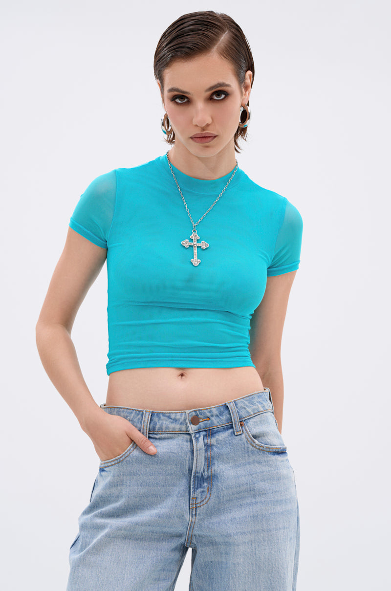 A woman with short, slicked-back hair wears the PAXTON DOUBLE LINED MESH SHORT SLEEVE TEE IN TURQUOISE, paired with light blue jeans, hoop earrings, and a silver cross necklace. She stands with one hand in her pocket against a plain white background.