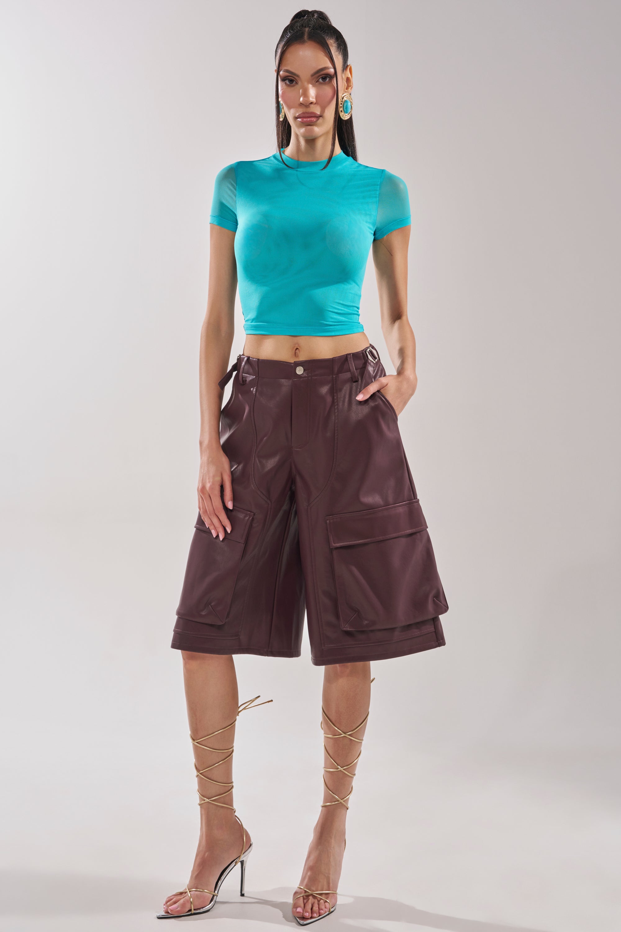 A woman poses against a plain background wearing the PAXTON Double Lined Mesh Short Sleeve Tee in turquoise, high-waisted brown knee-length cargo shorts, strappy gold heels, and statement earrings. 5