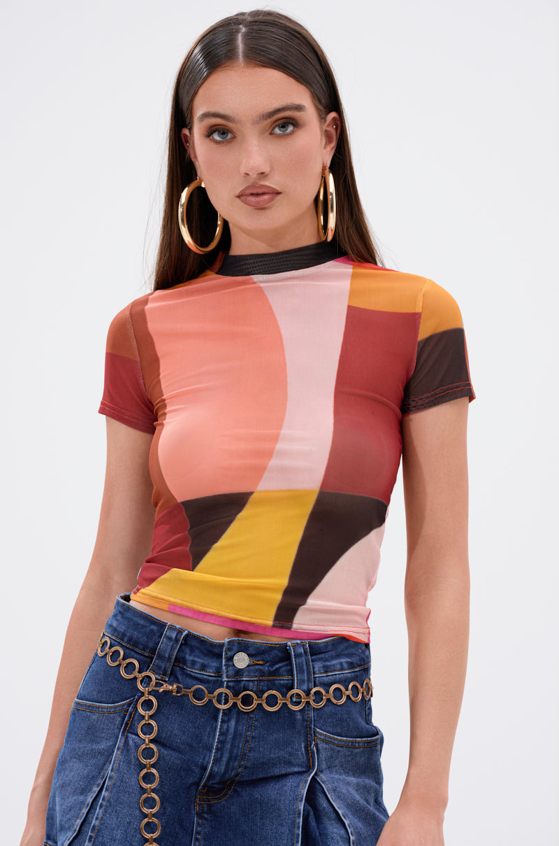 A woman with long brown hair wears the ABSTRACT THOUGHTS MESH SHORT SLEEVE TEE, featuring a stretchy, semi-sheer multicolored abstract print. She pairs it with hoop earrings, high-waisted blue jeans, and a gold chain belt against a white background.