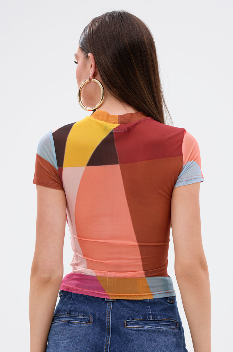 A woman with long brown hair, wearing large hoop earrings and the ABSTRACT THOUGHTS MESH SHORT SLEEVE TEE with a colorful abstract print, paired with blue jeans, is shown from the back against a plain background.
