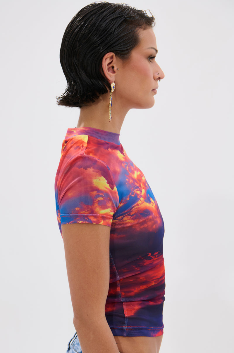 A person with short, slicked-back dark hair and a dangling earring poses in profile against a plain light background, wearing the HEAD IN THE CLOUDS MESH SHORT SLEEVE TEE with a vibrant sunset cloud photorealistic print. 3