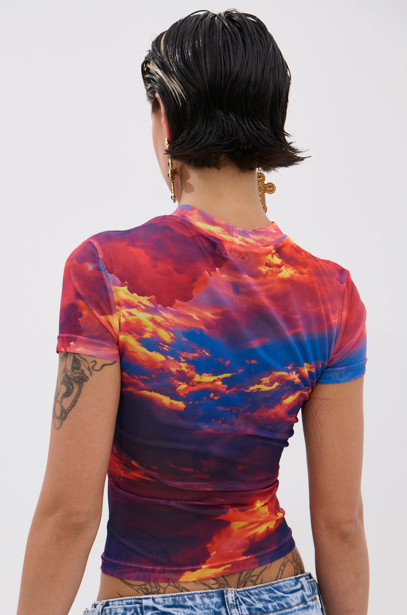 A person with short, dark wet-look hair is shown from the back wearing the HEAD IN THE CLOUDS MESH SHORT SLEEVE TEE featuring a vibrant sunset graphic, paired with light blue jeans, visible arm tattoos, and a dangling earring. 4