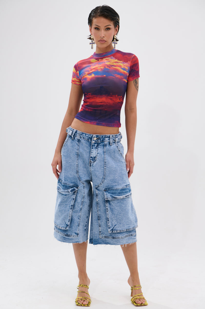 Person stands against a white background wearing the HEAD IN THE CLOUDS MESH SHORT SLEEVE TEE, featuring a vibrant graphic sunset print and curve-hugging fit, paired with wide knee-length denim shorts, yellow strappy heels, and statement earrings. 5