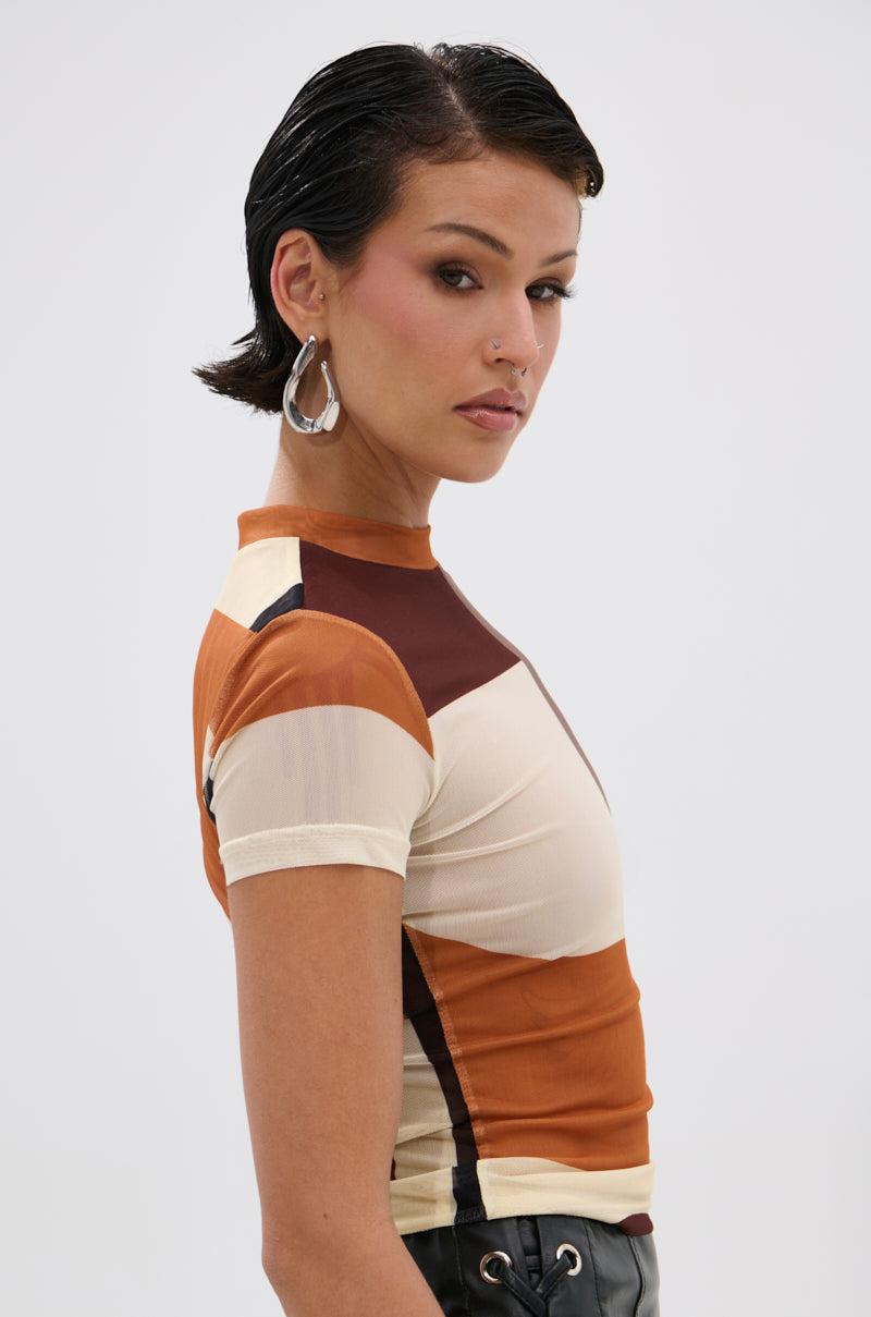 Woman with short, slicked-back dark hair and hoop earrings wears the SYMBOLISM MESH SHORT SLEEVE TEE with color-blocked beige, brown, and orange panels, glancing over her shoulder against a plain light background for a retro vibe.