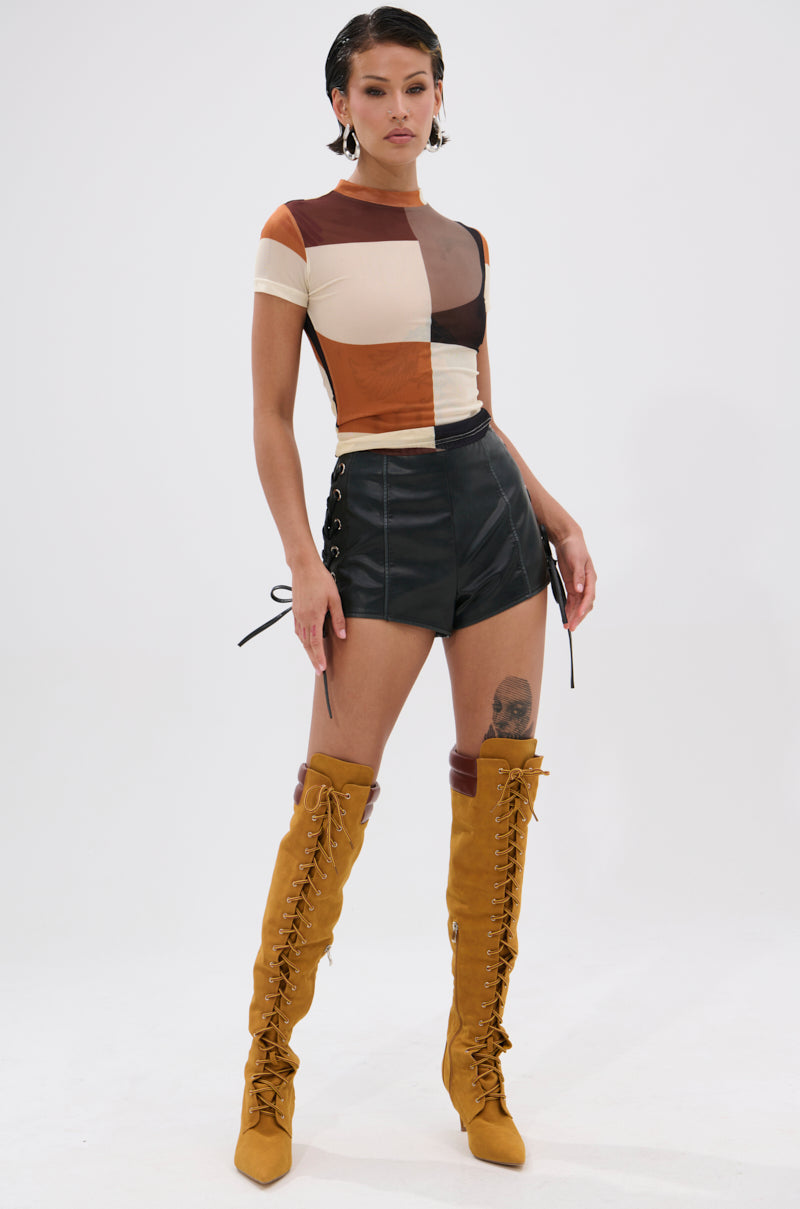 A woman with short dark hair, earrings, and a visible thigh tattoo poses confidently against a white background in the SYMBOLISM MESH SHORT SLEEVE TEE featuring a vintage-inspired abstract print, paired with black lace-up shorts and mustard yellow thigh-high boots.