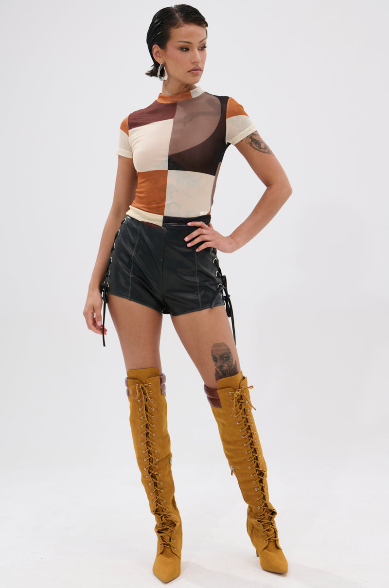 A person poses confidently in the SYMBOLISM MESH SHORT SLEEVE TEE, black leather lace-up shorts, and tall mustard-yellow lace-up boots against a plain white background.
