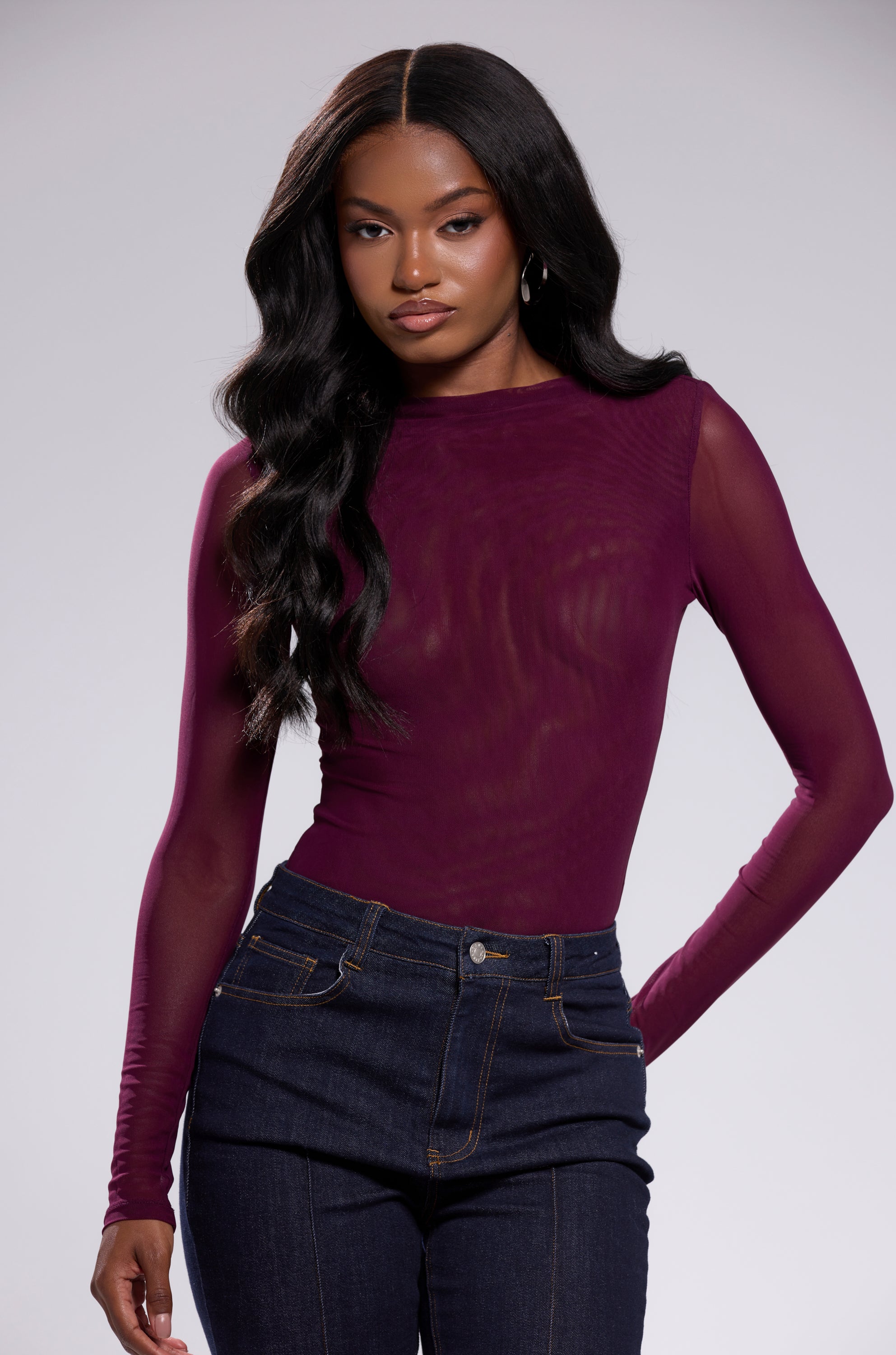 A woman with long, wavy black hair confidently models the PAXTON Sexy Mesh Long Sleeve Bodysuit in plum purple with high-waisted dark blue jeans against a plain light background.