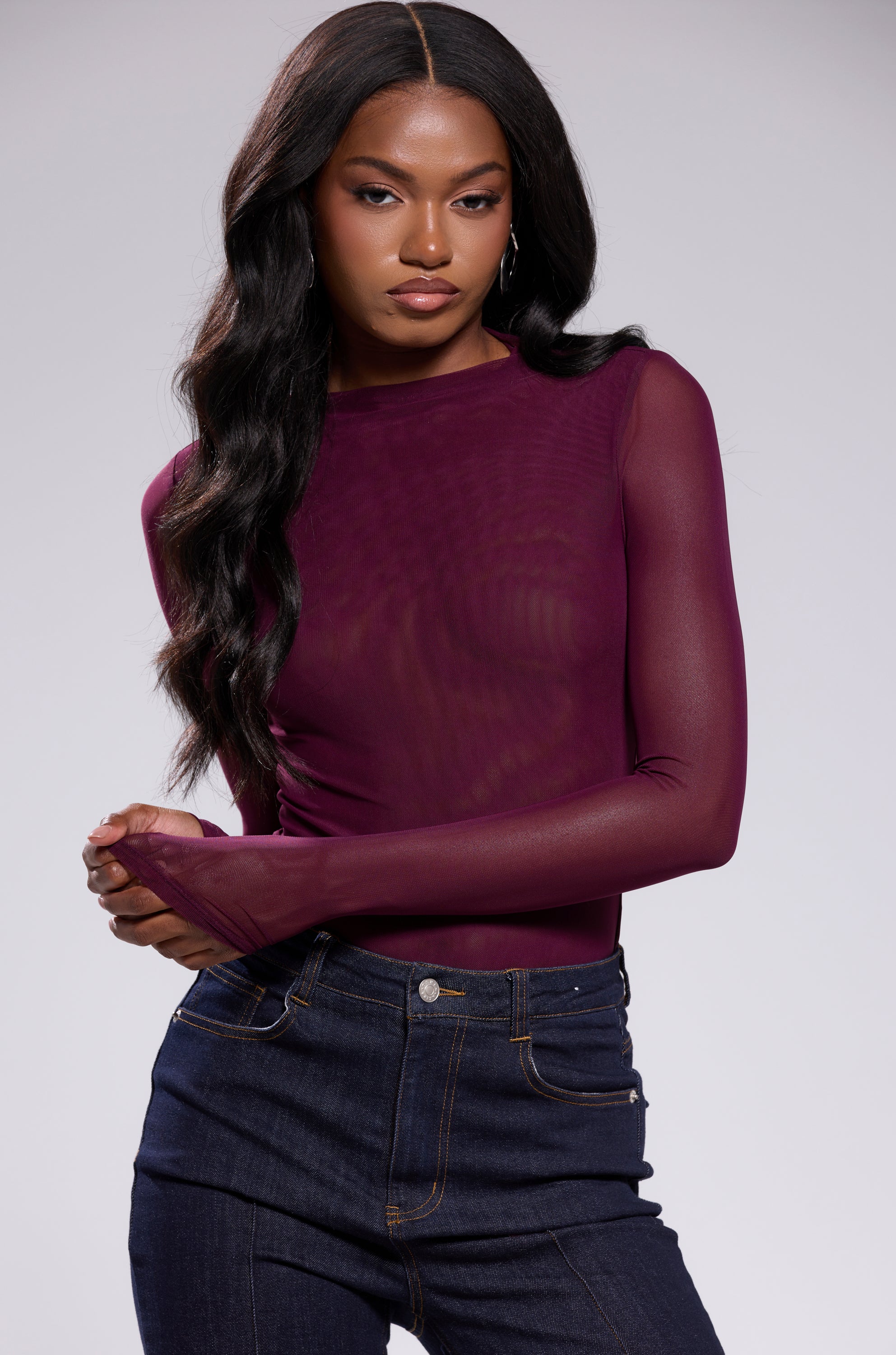 A woman with long, wavy dark hair wears the Paxton Sexy Mesh Long Sleeve Bodysuit in plum purple with high-waisted dark jeans, standing confidently against a light gray background.
