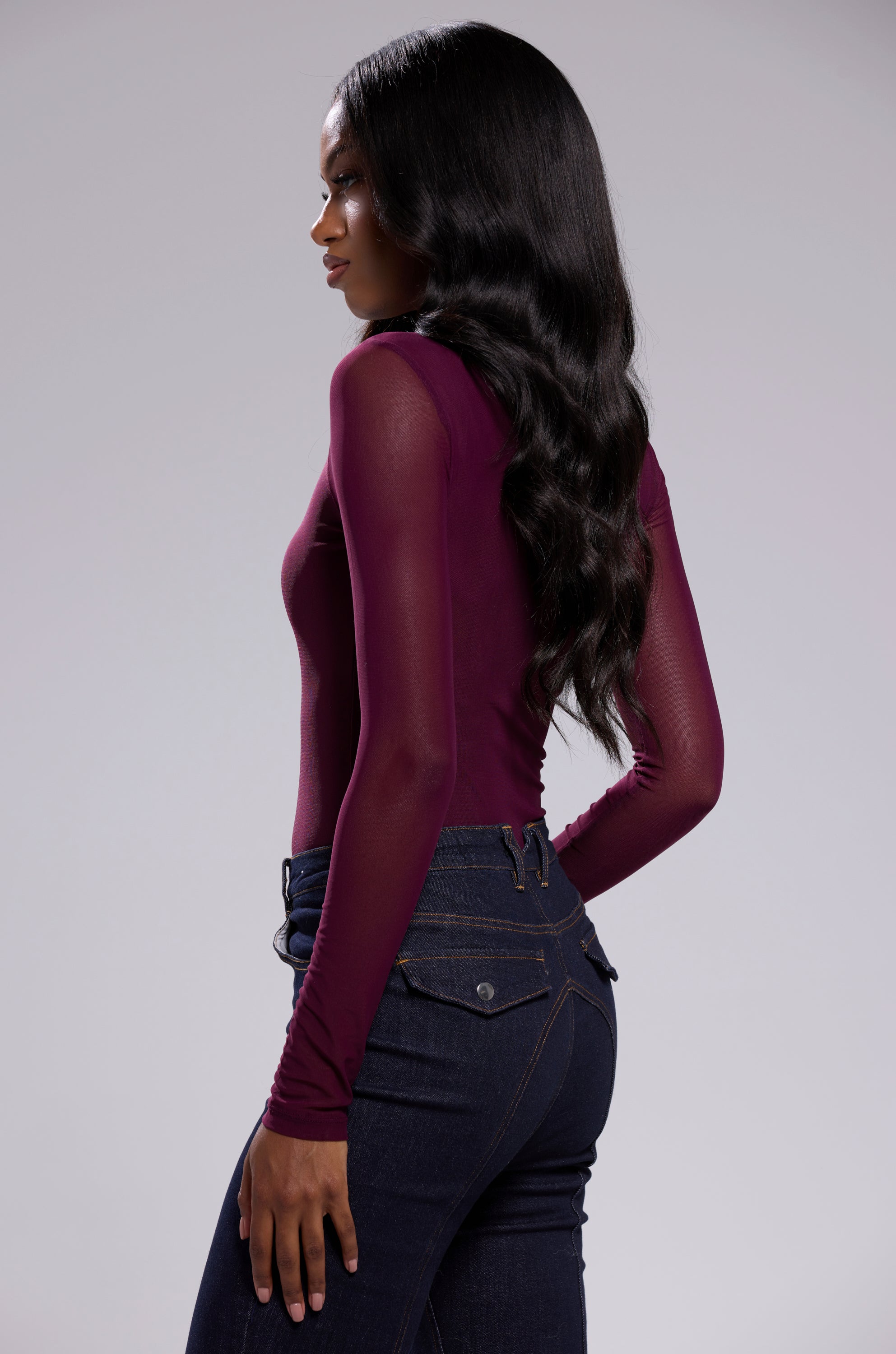 A woman with long, wavy black hair in profile wears the PAXTON SEXY MESH LONG SLEEVE BODYSUIT IN PLUM PURPLE and dark blue high-waisted jeans against a plain light gray background.