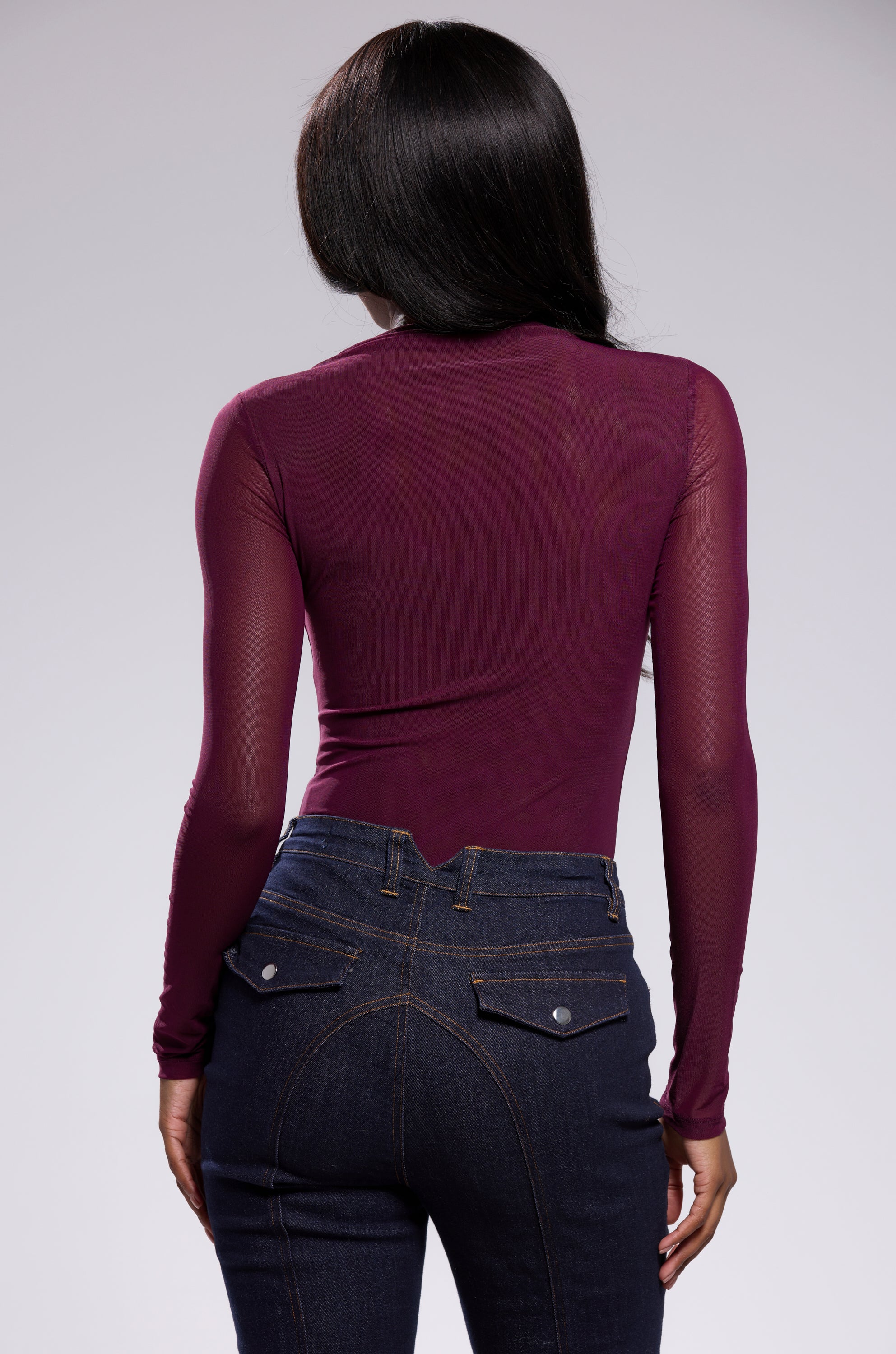 A person with long dark hair, seen from behind, models the PAXTON Sexy Mesh Long Sleeve Bodysuit in plum purple with high-waisted dark blue jeans against a plain light background.