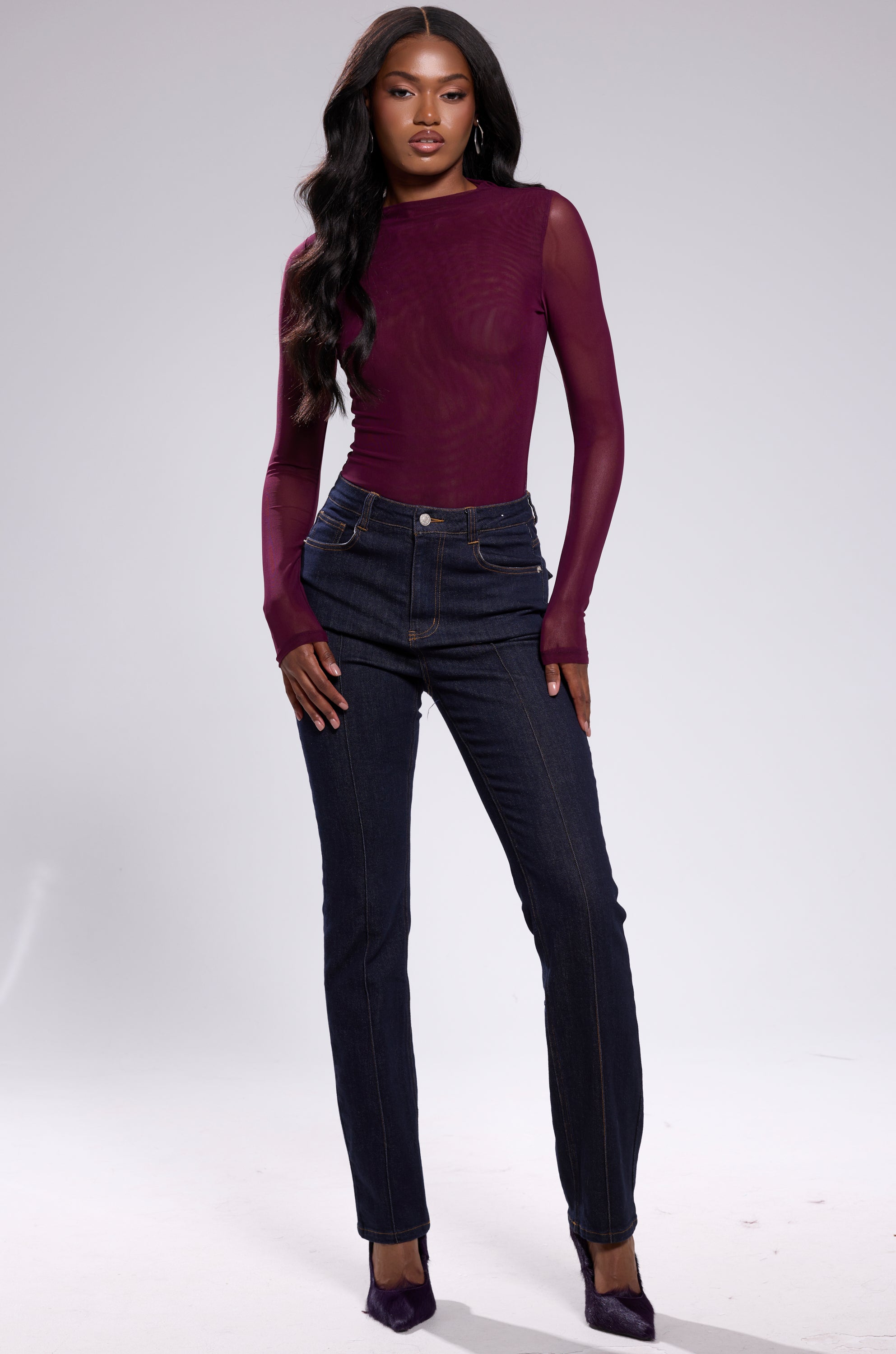 A woman faces forward wearing the PAXTON Sexy Mesh Long Sleeve Bodysuit in plum purple, styled with dark blue high-waisted jeans and burgundy heels, her long wavy hair flowing against a neutral background.