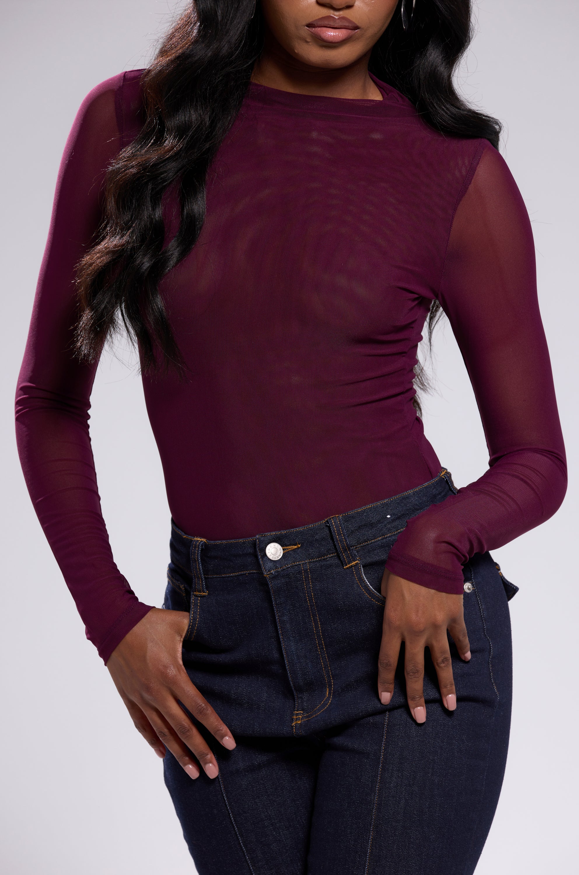 A woman wears the PAXTON Sexy Mesh Long Sleeve Bodysuit in Plum Purple with high-waisted dark blue jeans, standing against a plain background, one hand on her hip and her face partially out of frame.