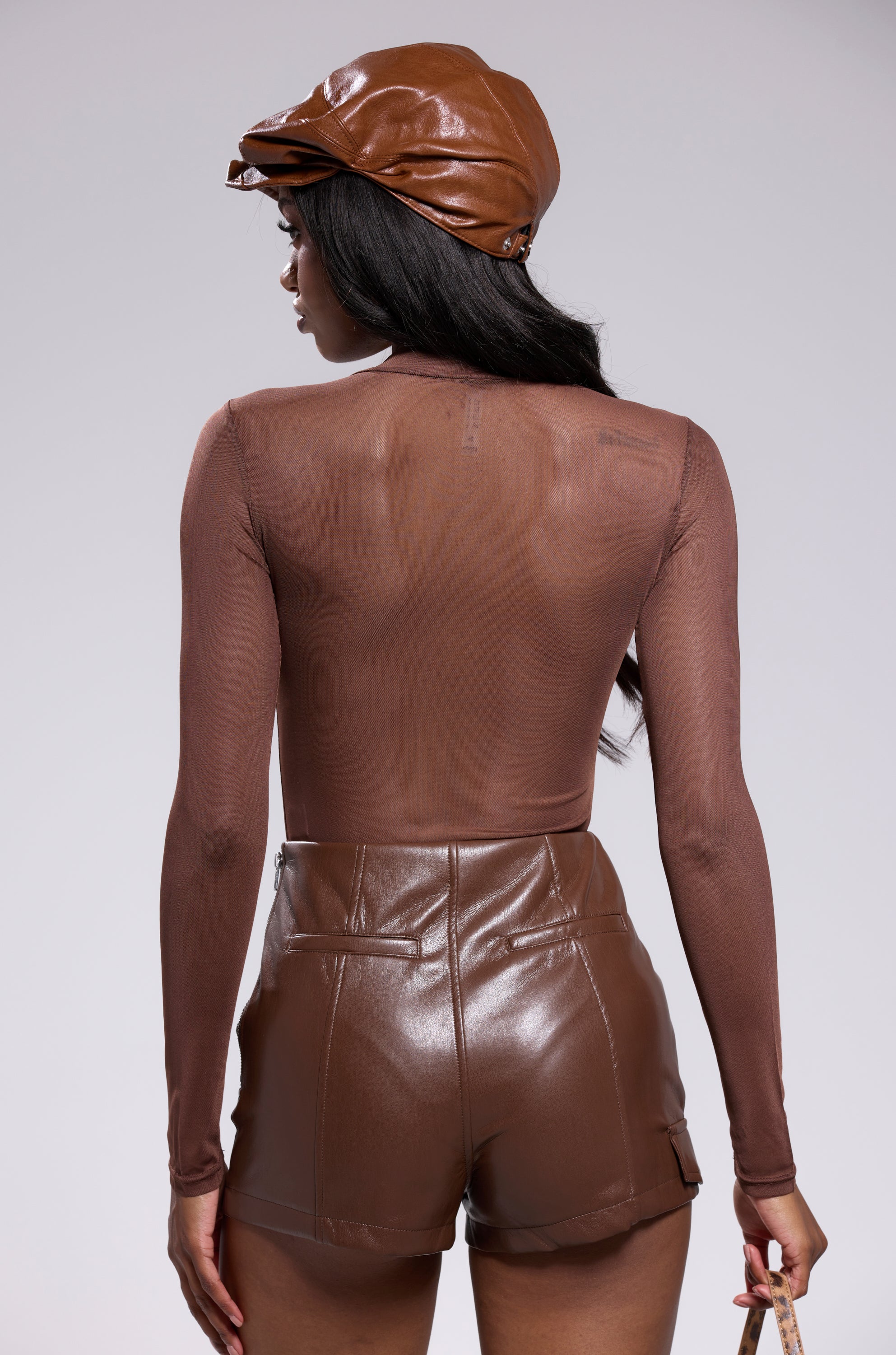 A woman faces away, wearing the PAXTON Sexy Mesh Casual Bodysuit in Brown, paired with high-waisted brown leather shorts and a matching cap, against a plain light background.