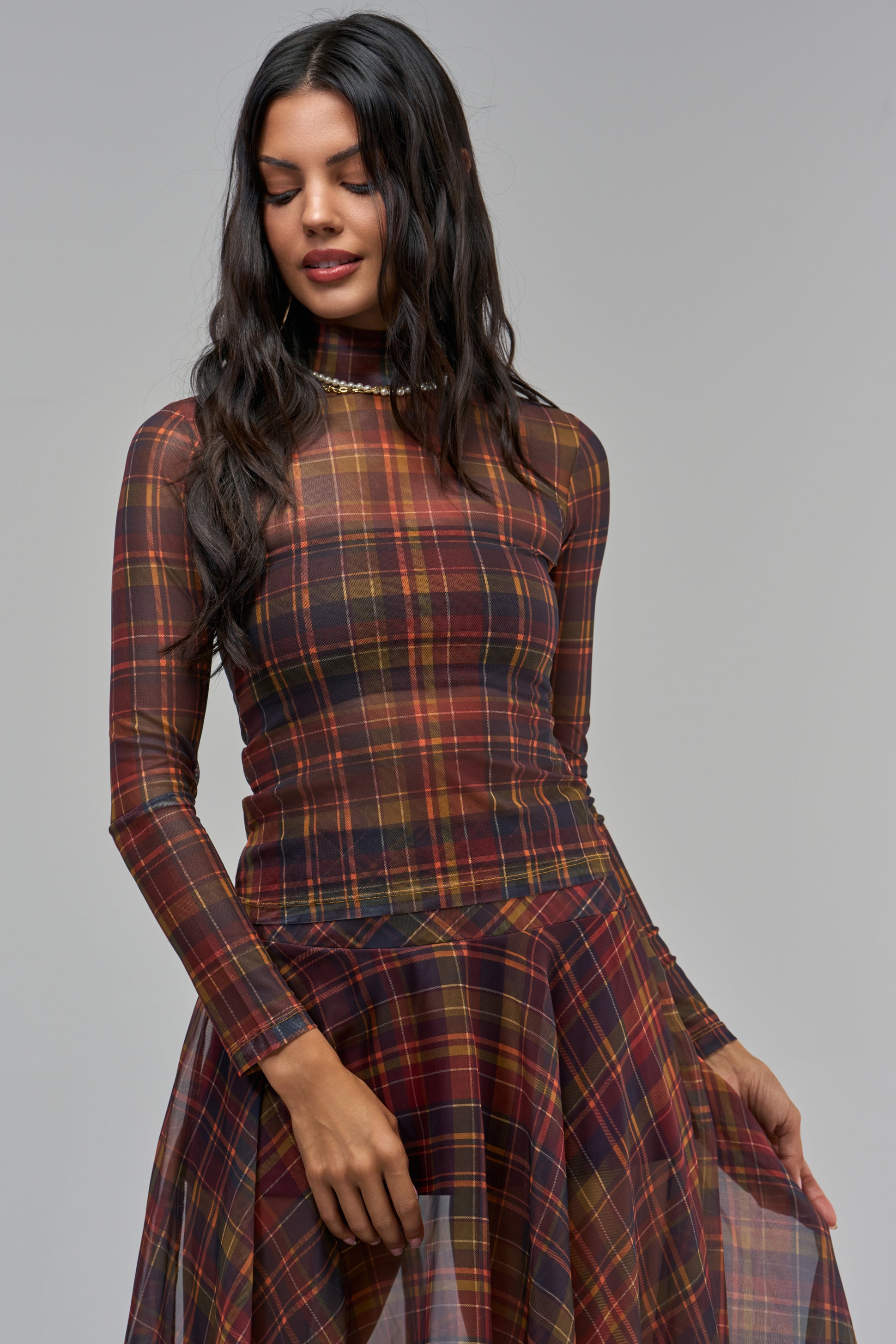 A person wears the PRETTY IN PLAID MESH MOCK NECK TOP and a matching skirt in a brown and red plaid mesh fabric, perfect for fall layering.