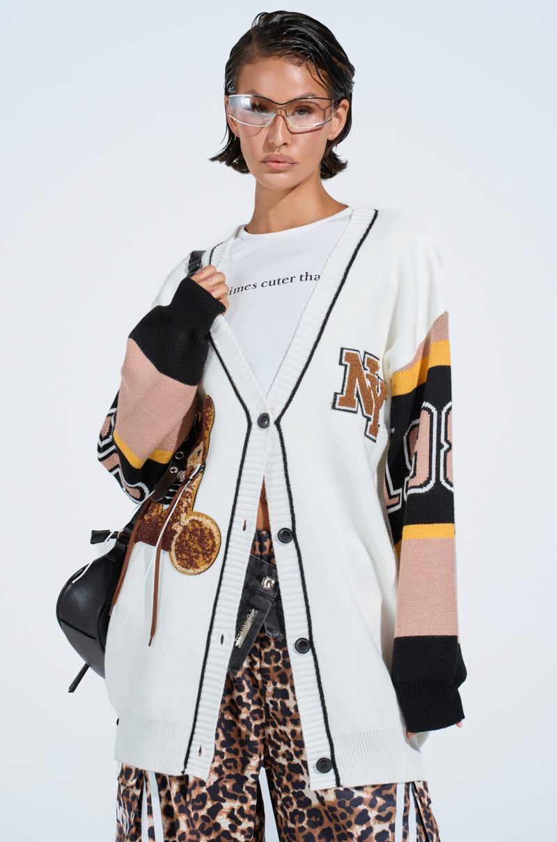 A woman wears the COZY AS CAN BE CARDIGAN with black trim and colorful sleeves over a white T-shirt, paired with leopard print pants. She has clear glasses, carries a black shoulder bag, and has her hair slicked back.