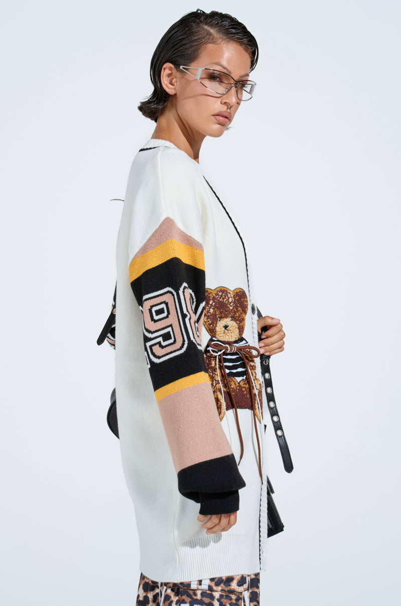 A person with short dark hair and glasses wears the COZY AS CAN BE CARDIGAN, featuring black, yellow, and tan stripes, a teddy bear patch design, and the number 99 on the sleeve, standing against a plain light background.