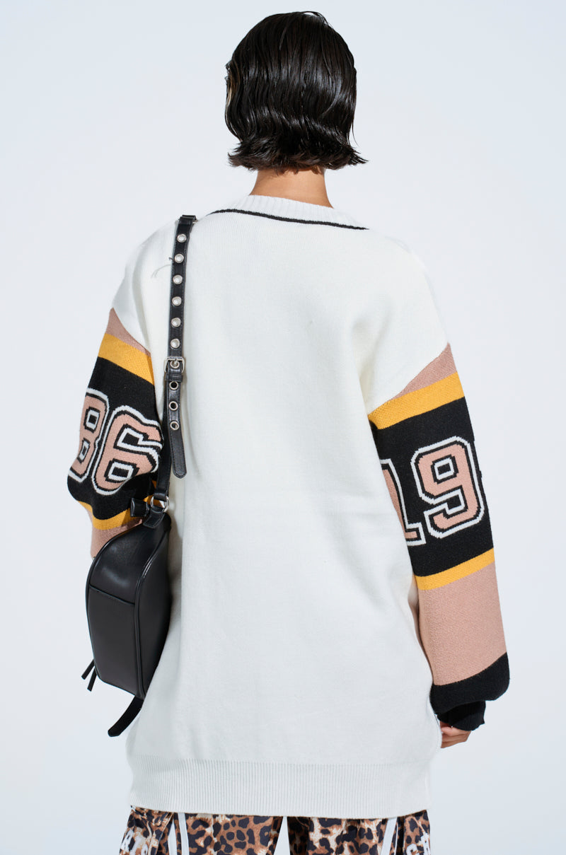 A person with short dark hair, seen from behind, wears the COZY AS CAN BE CARDIGAN—an oversized white sweater with "96" and "19" on the sleeves—paired with a black shoulder bag and leopard print pants.