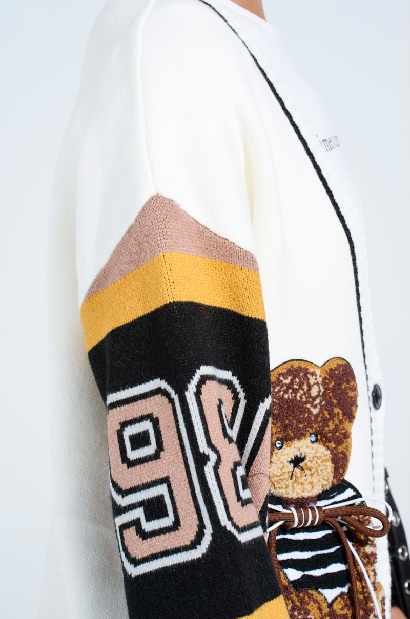 A close-up of the COZY AS CAN BE CARDIGAN, an oversized sweater with a large 96 patch, brown and yellow striped sleeves, and a playful teddy bear patch in a striped shirt on the front.