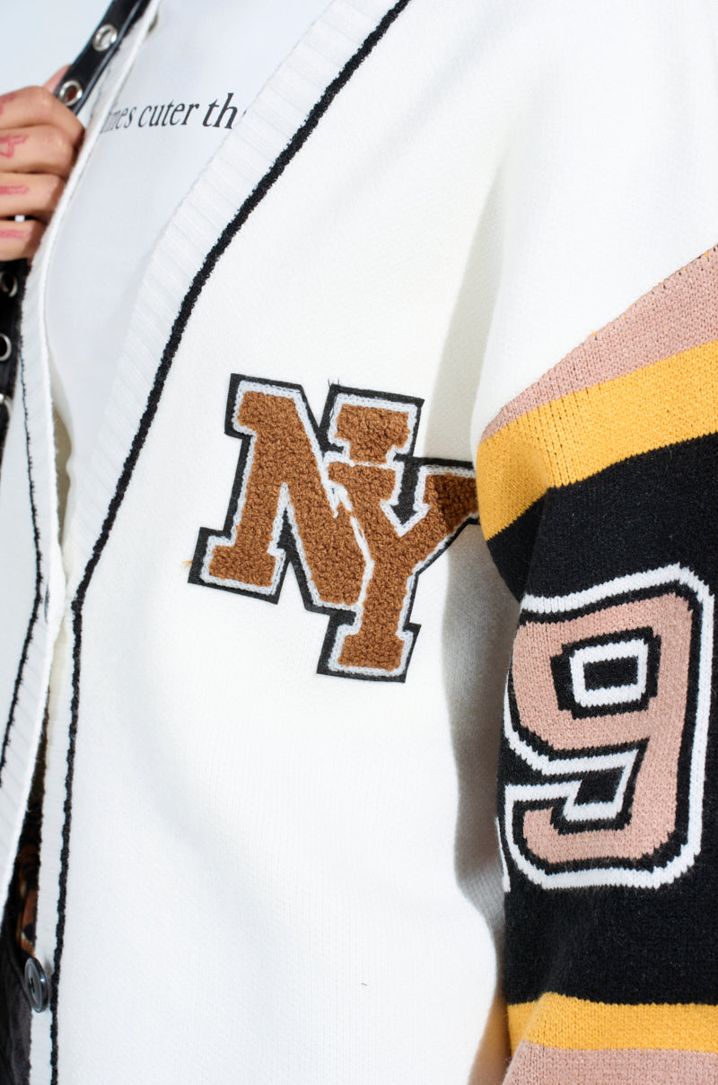 Close-up of a person wearing the COZY AS CAN BE CARDIGAN—an oversized sweater with black trim, a large brown "NY" patch, and a black sleeve featuring yellow and brown stripes plus the number "9." A printed shirt peeks out underneath.