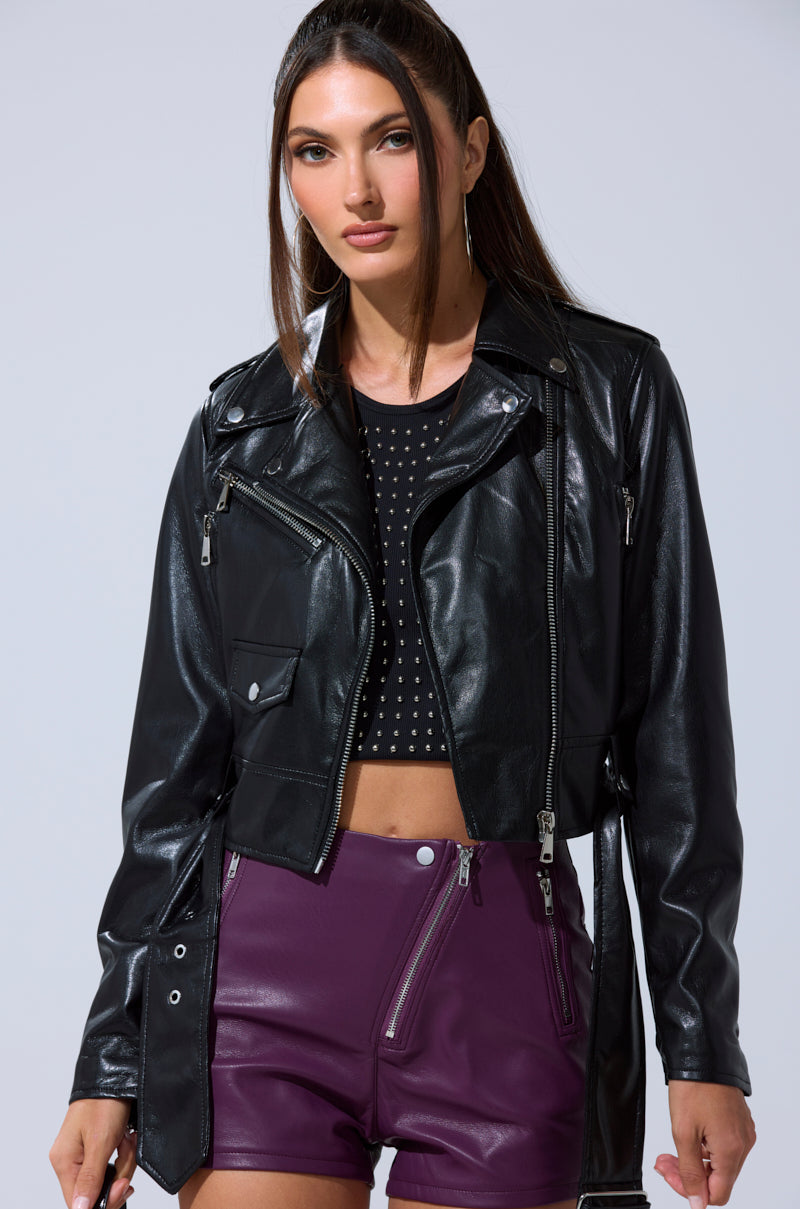 A woman with long brown hair wears the BUTTER LUXE EVERYDAY MOTO jacket over a black studded crop top and high-waisted purple leather shorts, standing against a plain light gray background.