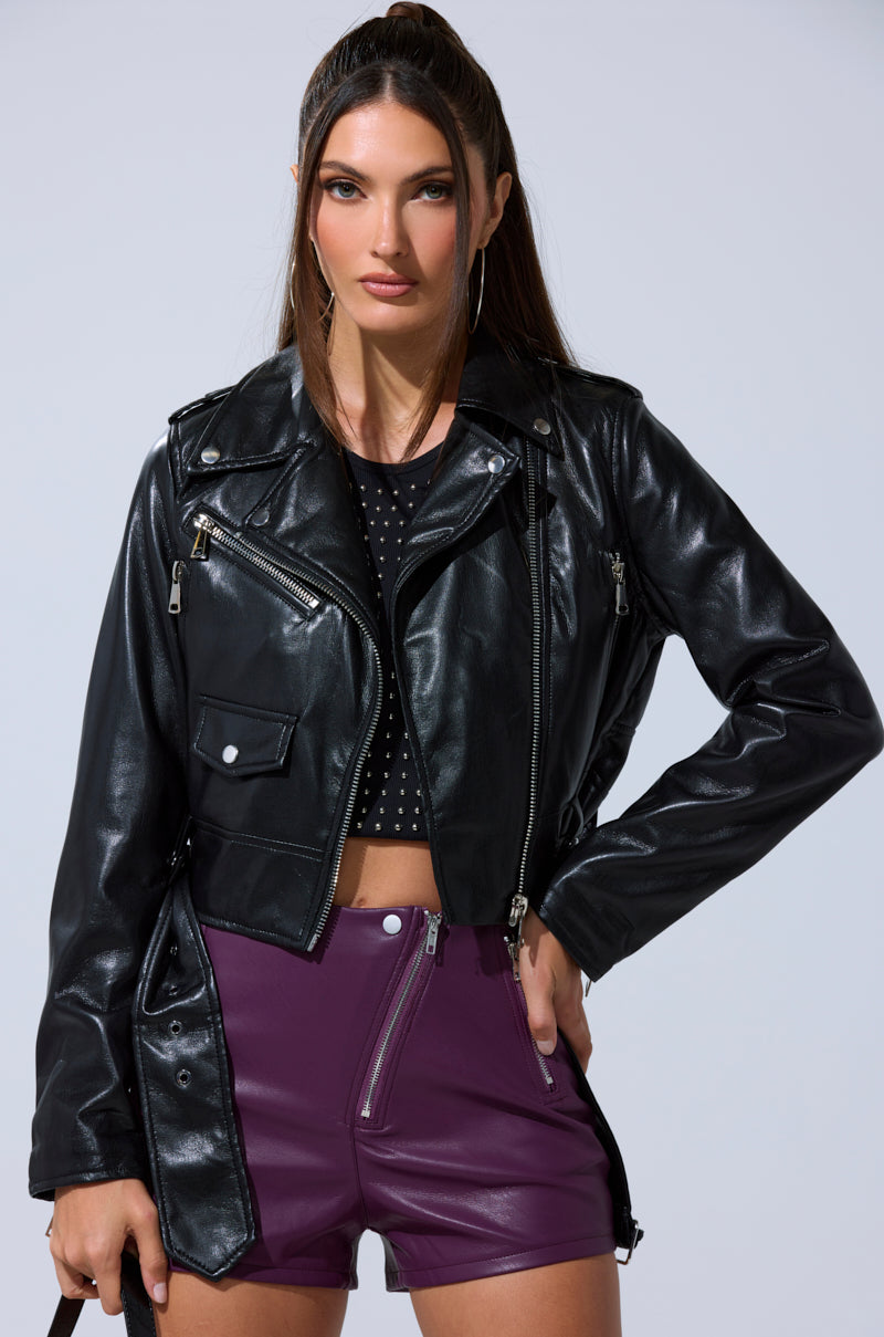 A woman with long brown hair wears the BUTTER LUXE EVERYDAY MOTO black faux leather jacket, paired with a black studded crop top and high-waisted purple shorts with front zippers, posing with one hand on her hip against a light gray background.