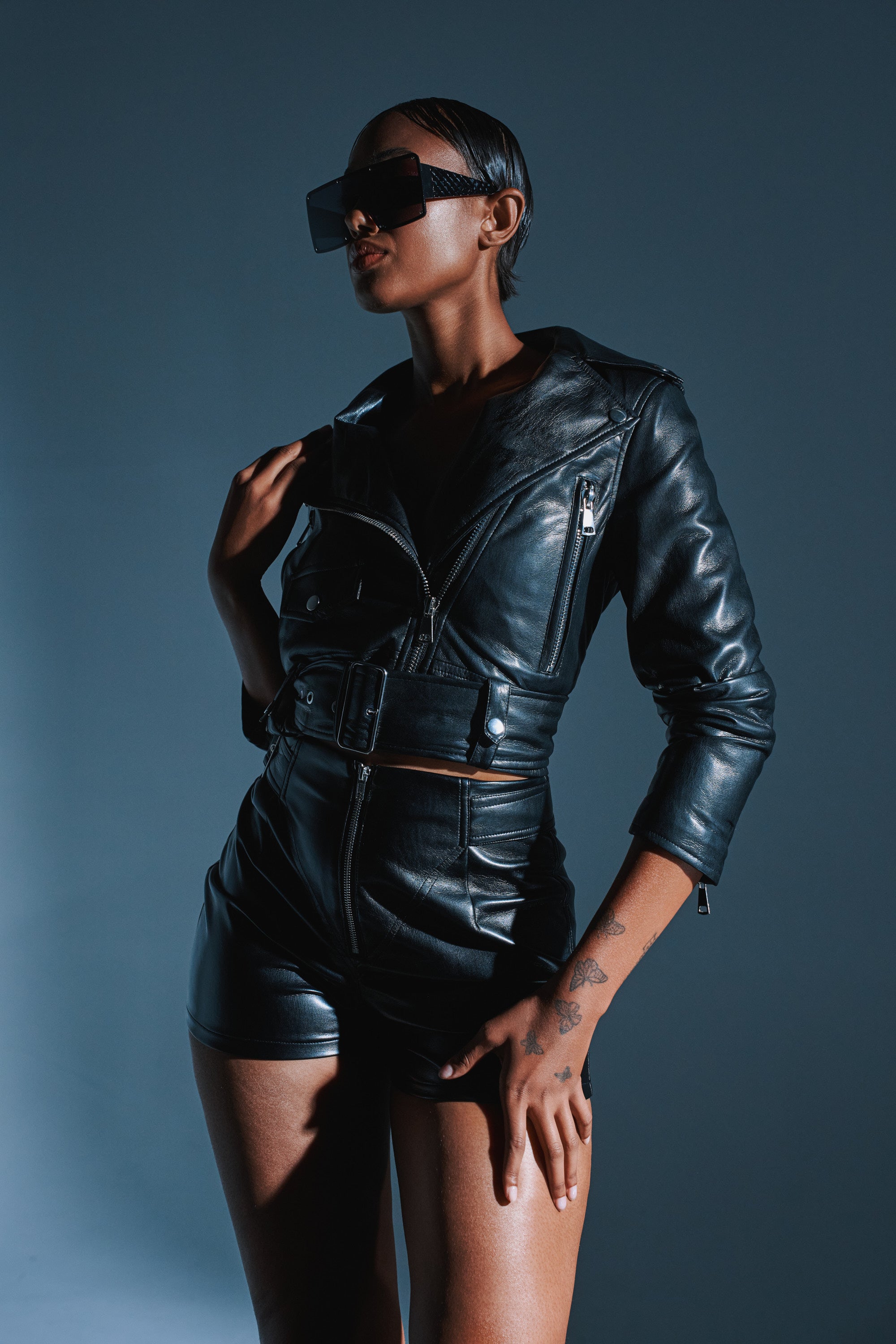 Against a dark background, a person with short hair and large sunglasses poses confidently in the BUTTER LUXE EVERYDAY MOTO jacket and shorts, one hand resting on their jacket collar.