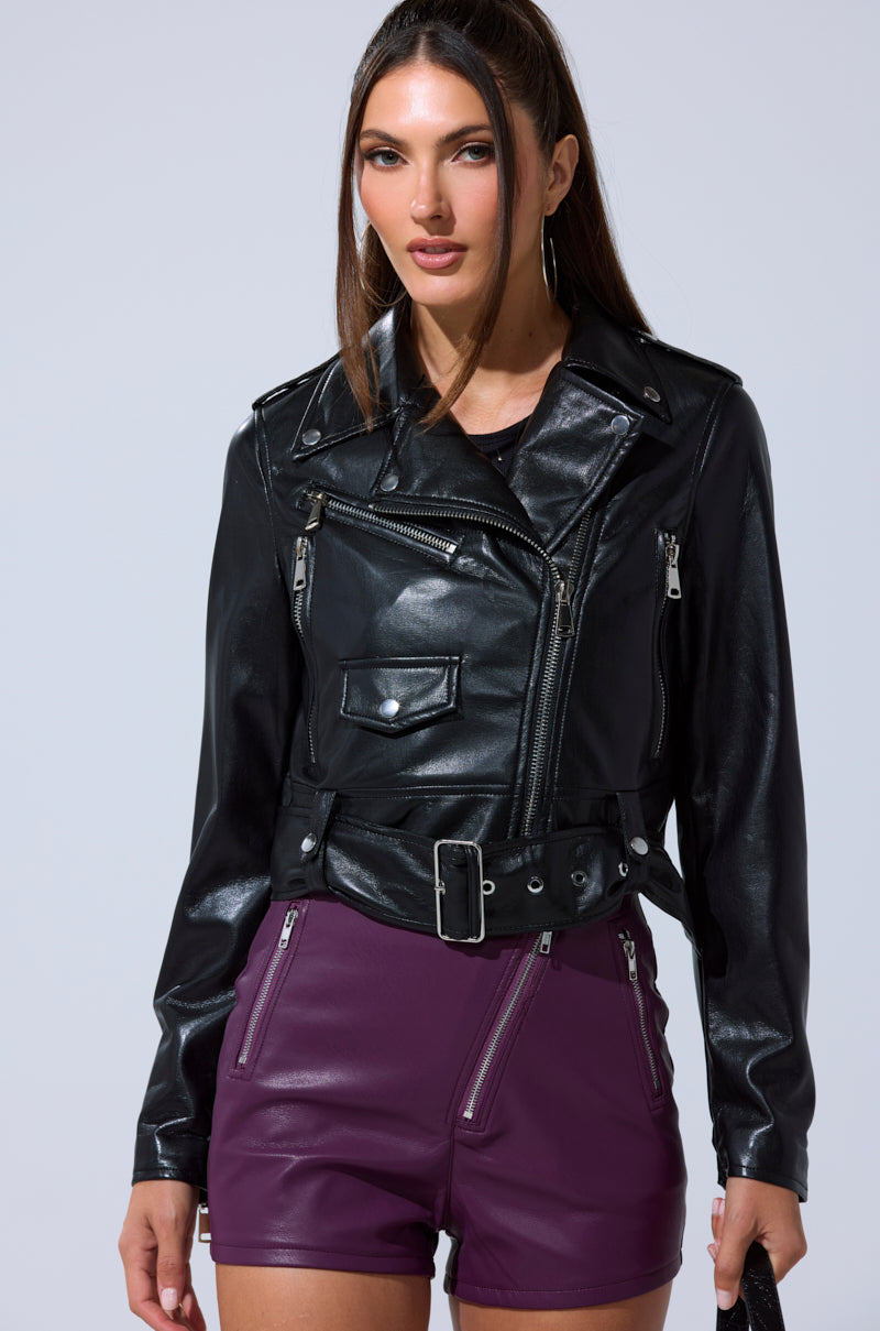 A woman wears the BUTTER LUXE EVERYDAY MOTO black faux leather jacket with silver zippers and buckles, paired with high-waisted purple shorts with zipper details, standing against a light gray background.