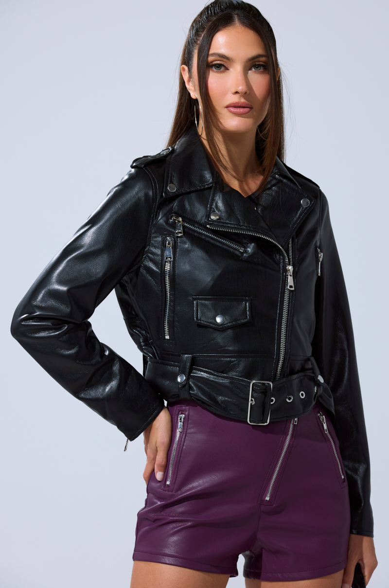 A woman with long brown hair wears the BUTTER LUXE EVERYDAY MOTO jacket and high-waisted purple shorts with zippered pockets, standing confidently against a light gray background.