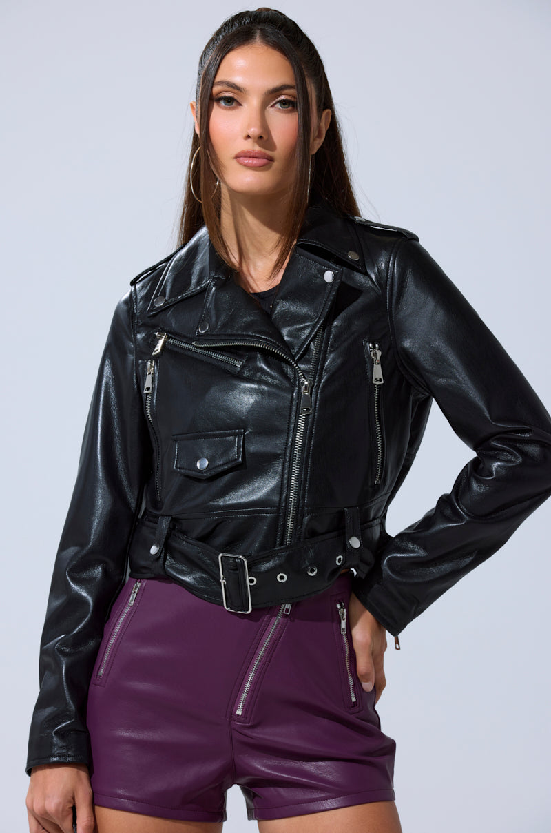 A woman with long brown hair wears the BUTTER LUXE EVERYDAY MOTO black faux leather biker jacket with silver zippers and buttons, paired with high-waisted purple shorts. She stands with one hand on her hip against a plain background.