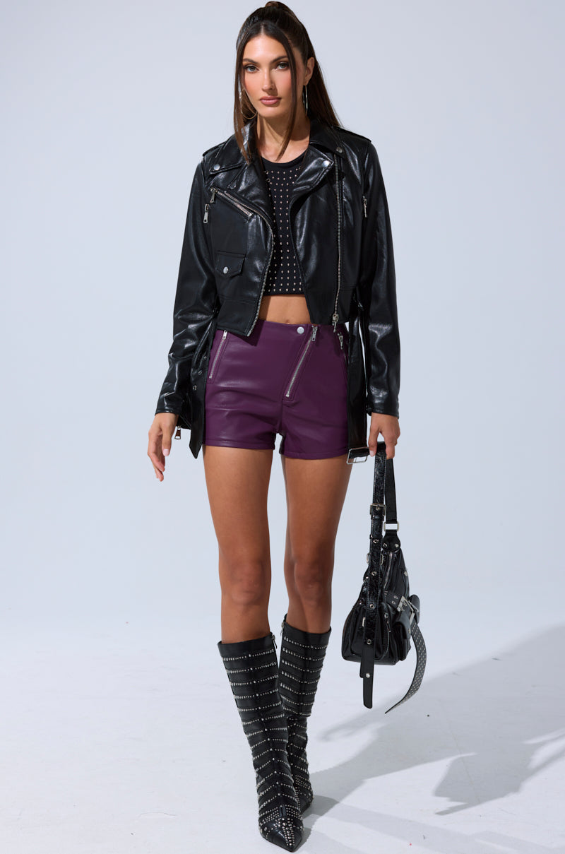 A woman stands confidently against a plain light background wearing the BUTTER LUXE EVERYDAY MOTO black faux leather biker jacket, paired with a black crop top, purple leather shorts, studded knee-high boots, and holding a black handbag.