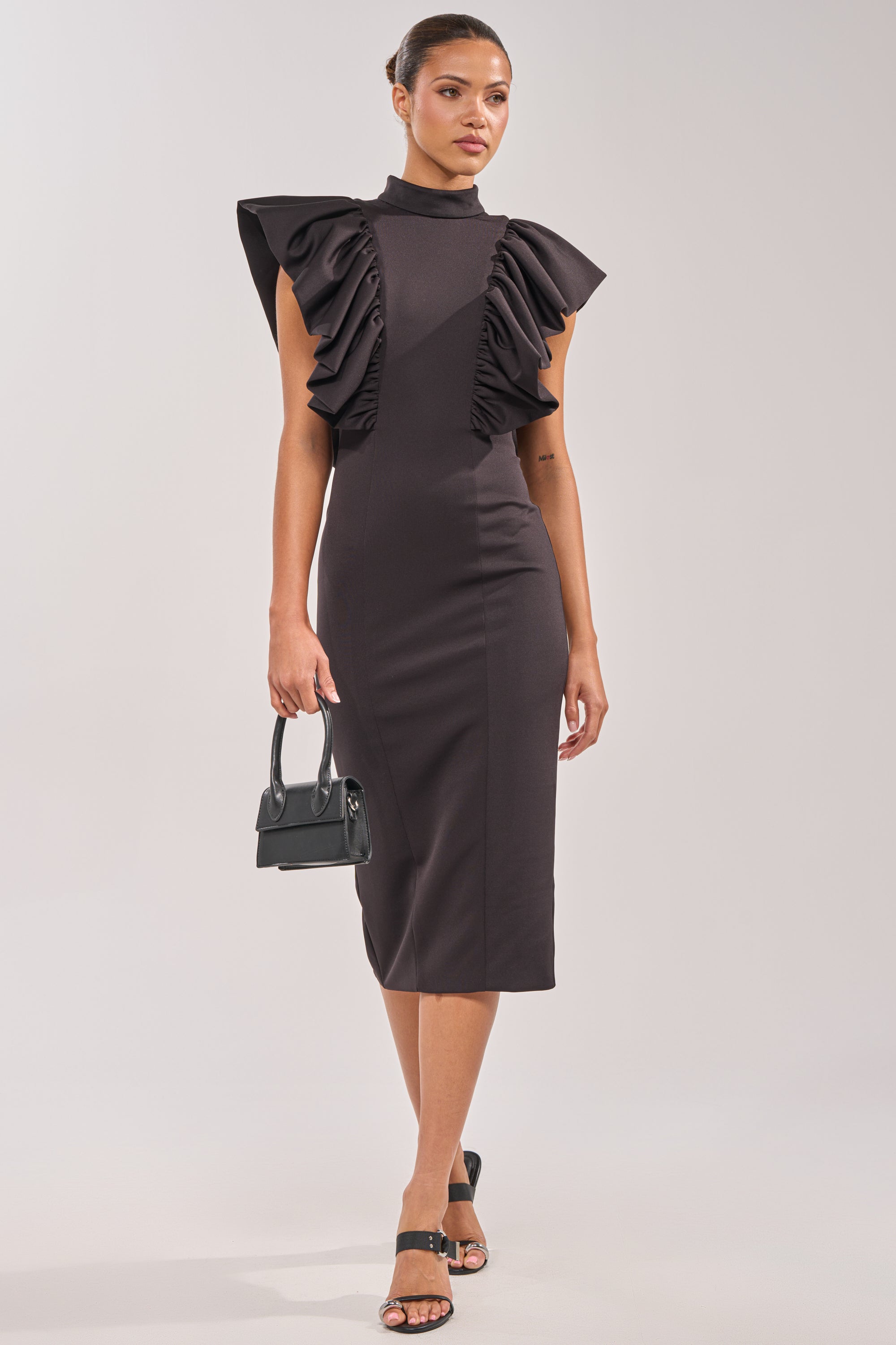 A woman wears the TRUTH BE TOLD MIDI DRESS—a fitted black ruffle scuba dress with dramatic sleeves—styled with black heeled sandals and a small handbag. Her sleek bun completes this chic look, ideal for evening events or as a wedding guest outfit. 0