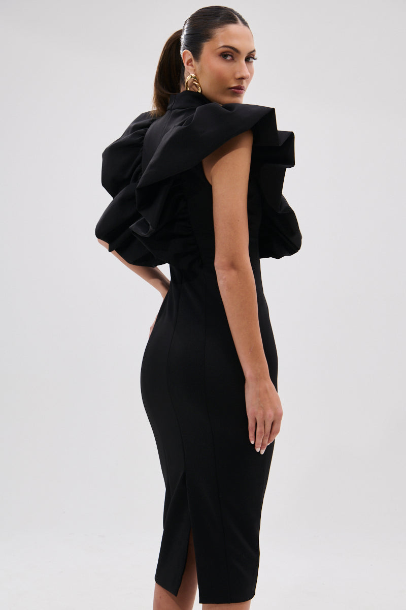 A woman stands sideways against a plain light background wearing the TRUTH BE TOLD MIDI DRESS, featuring a fitted sculptural silhouette and dramatic ruffle shoulders. Her hair is in a sleek ponytail and she accessorizes with large gold earrings. 7