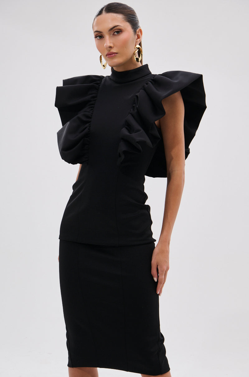 A woman poses against a plain light background, wearing the TRUTH BE TOLD MIDI DRESS—fitted and black with sculptural ruffle shoulders—styled with gold hoop earrings and a sleek hairstyle. 8