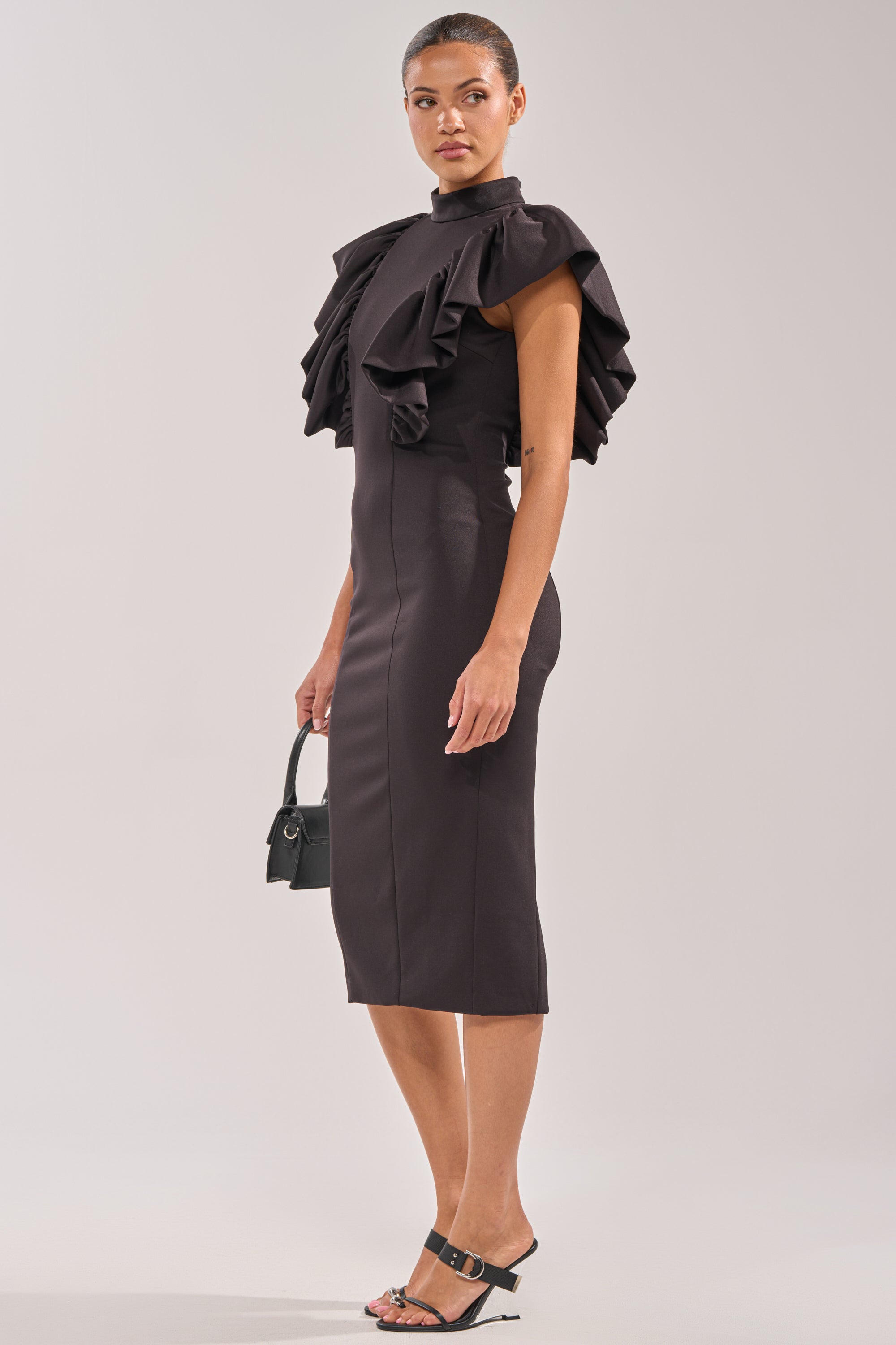 A woman in the TRUTH BE TOLD MIDI DRESS, featuring black ruffles and dramatic sleeves, poses sideways with a small black handbag and black heeled sandals. Her hair is pulled back against a plain light background. 2