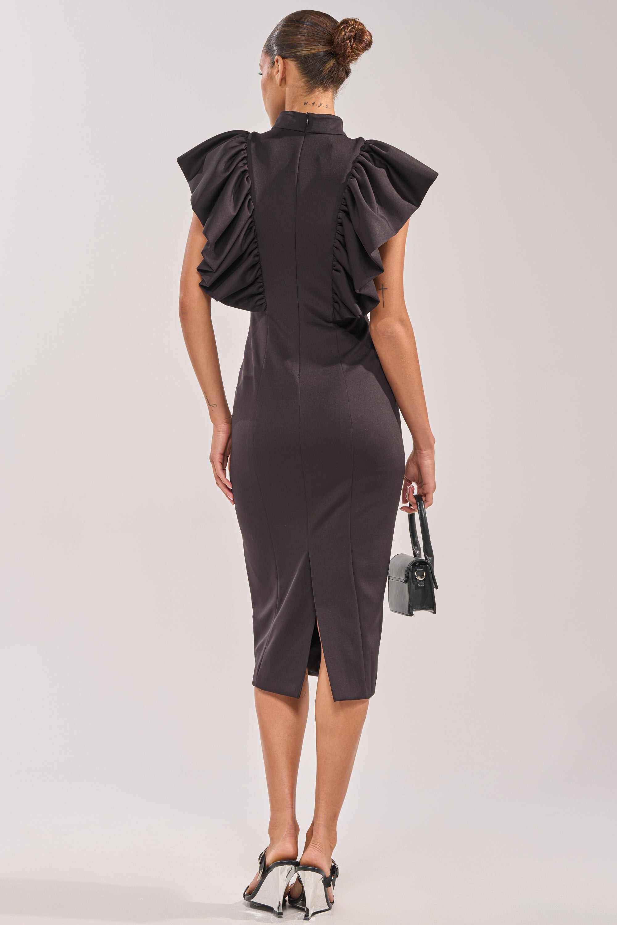 A woman faces away, wearing the TRUTH BE TOLD MIDI DRESS—a black ruffle midi with a fitted, knee-length silhouette and ruffled sleeves. She carries a small black handbag and wears two-tone bow-back heels, hair in a sleek bun. 3
