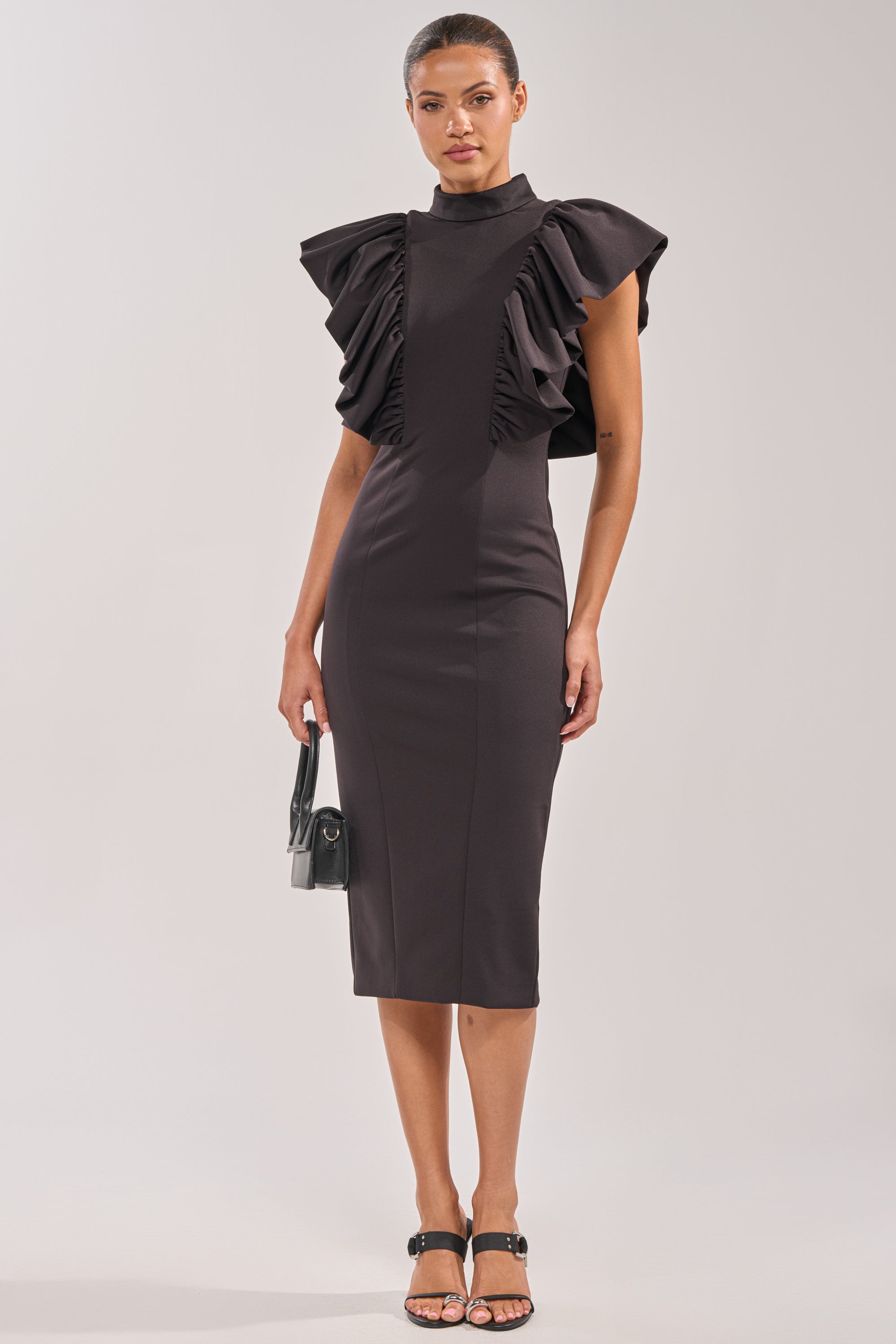 A woman models the TRUTH BE TOLD MIDI DRESS, a fitted black ruffle midi with dramatic sleeves, styled with a small black handbag and heeled sandals—ideal evening wear set against a simple light background. 4