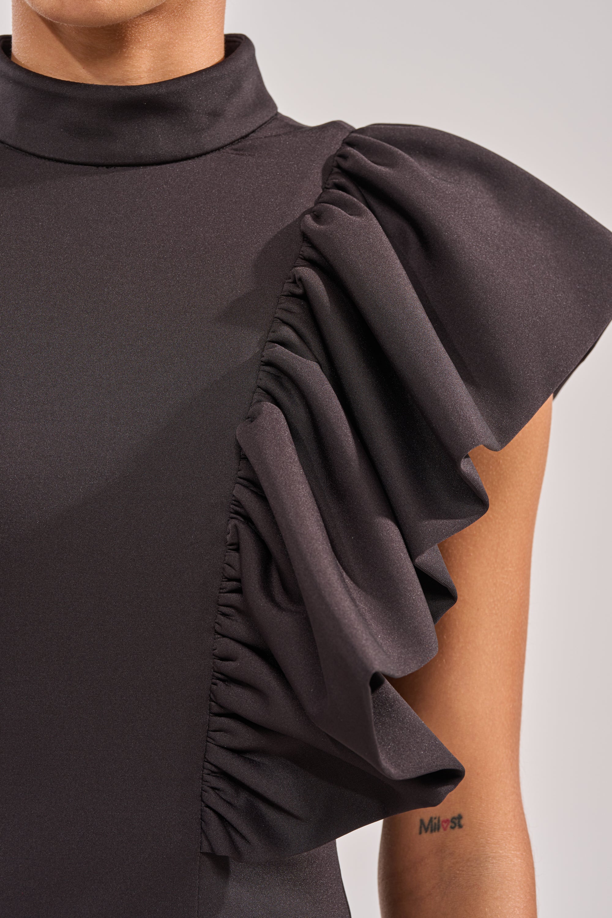 Close-up of a person in the TRUTH BE TOLD MIDI DRESS—black with a high neck and bold ruffled sleeve on one side. Elegant choice for evening events, with a glimpse of their arm revealing a small wrist tattoo. 5