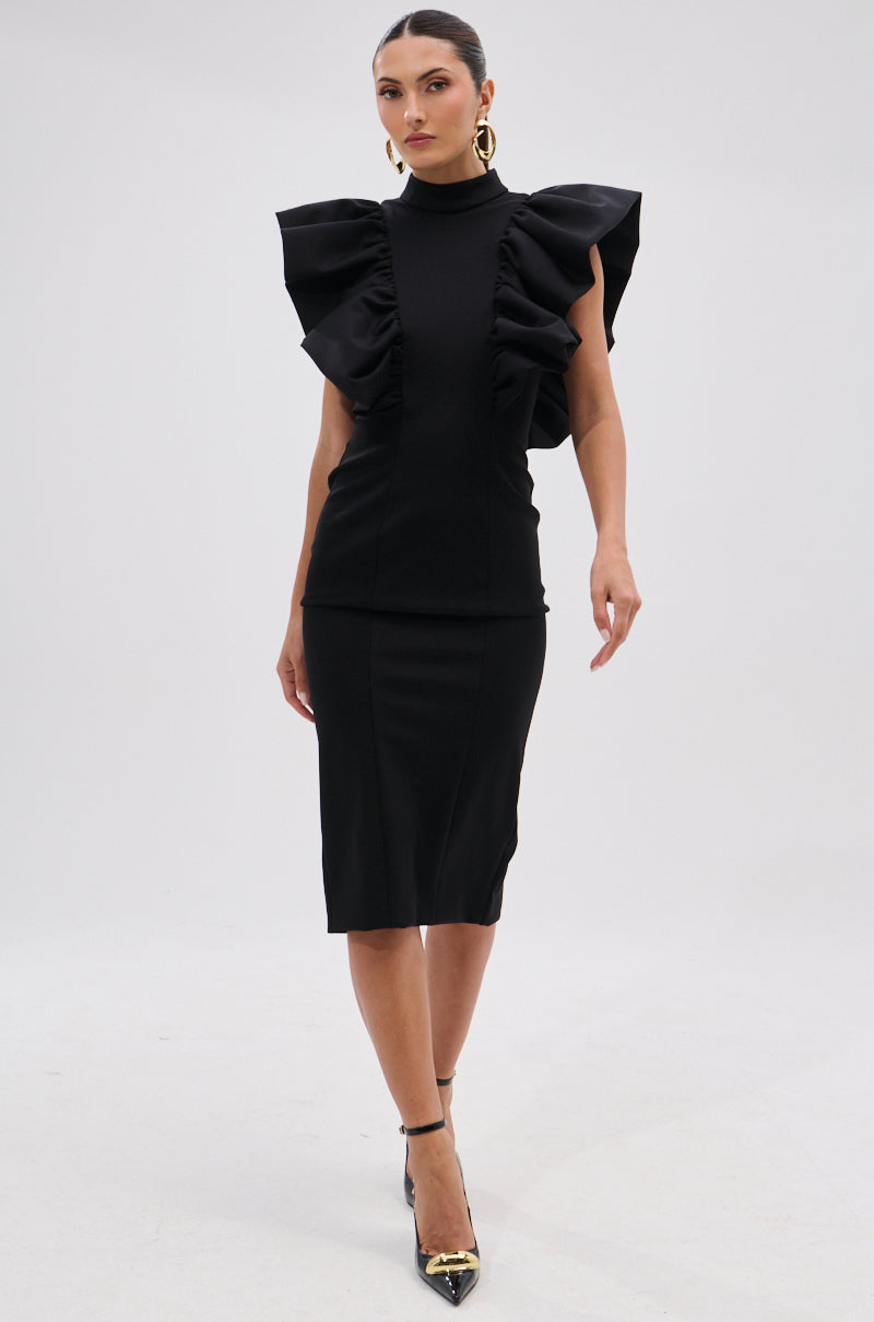 A woman walks confidently in the TRUTH BE TOLD MIDI DRESS, featuring sculptural ruffle shoulders, paired with black heels and gold hoop earrings against a plain light background. 13