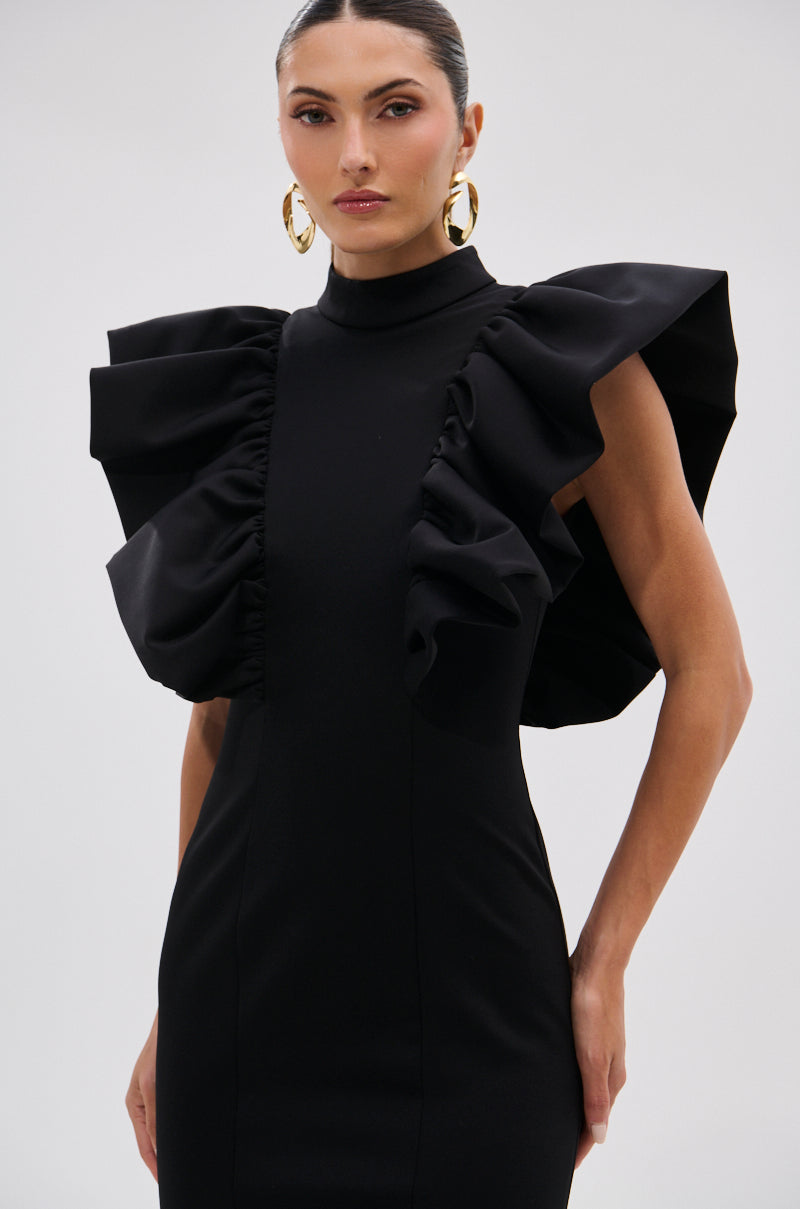 A woman wears the TRUTH BE TOLD MIDI DRESS—a fitted black midi dress with dramatic ruffle shoulders and a sculptural silhouette. She pairs it with slicked-back hair, large gold hoop earrings, and a confident expression against a plain background. 14