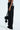 A person in DYLAN LACE DETAIL wide-leg denim with a small white detail on the back pocket holds a black handbag. The high-rise fit highlights the lace detail, adding elegance to their style. They're wearing white sneakers against a plain white background.