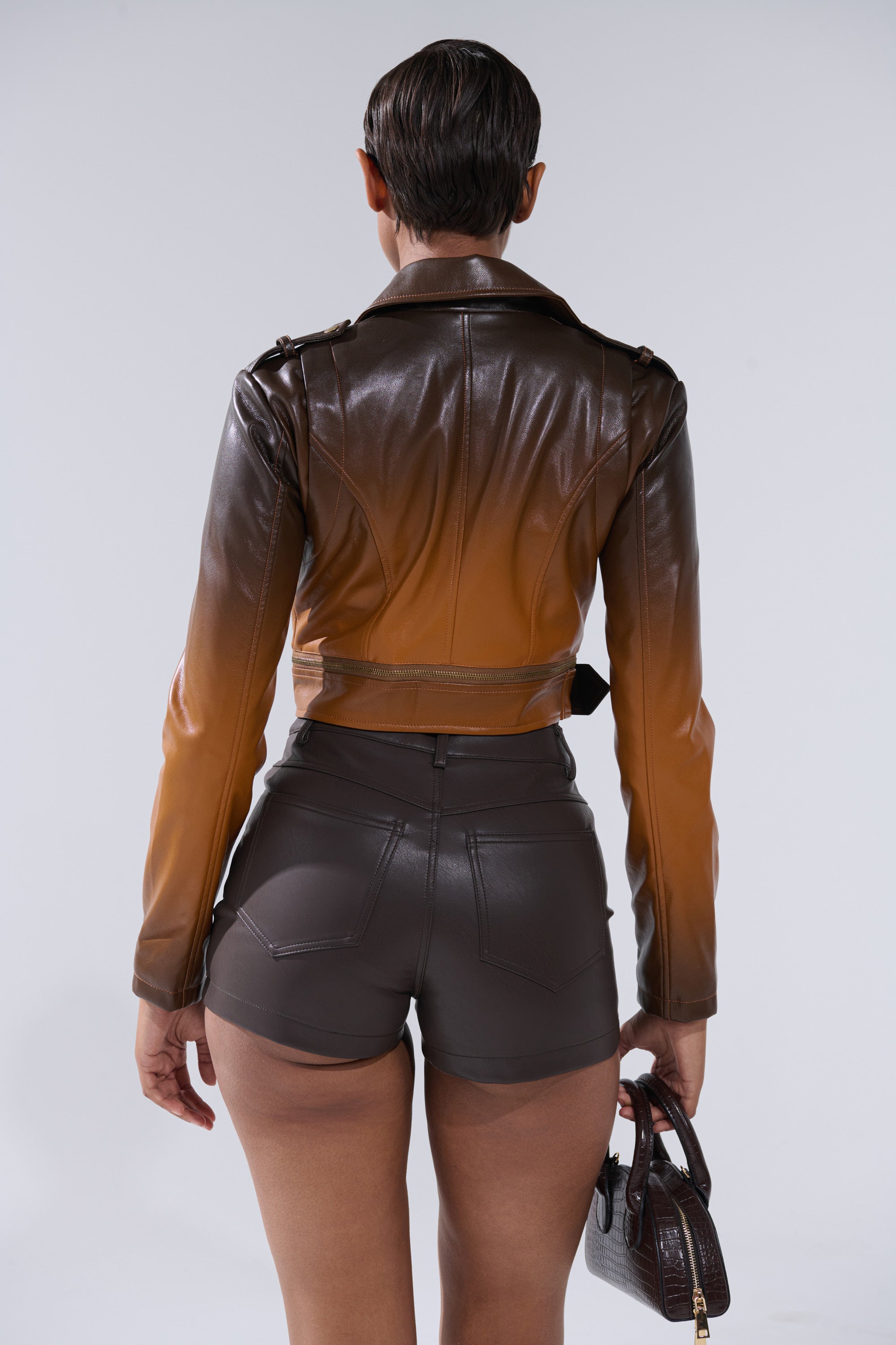 A person with short, slicked-back hair is seen from behind wearing the ENORA OMBRE MOTO jacket and matching shorts, holding a small brown handbag in their left hand against a plain background. 6